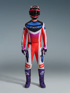 Motocross rider wearing complete racing gear, Alpinestars 2026 Techstar Knif Pants, Fluo orange and white design with blue accents, matching multi-colored jersey, purple boots, black and purple gloves, coordinated professional outfit for motocross racing
