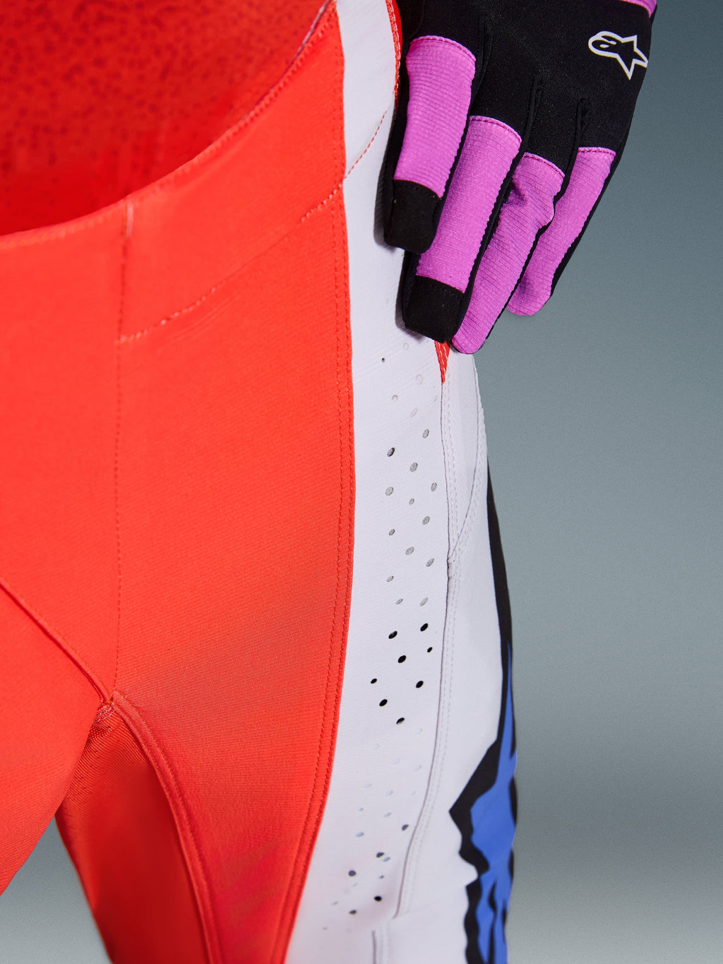 Alpinestars 2026 Techstar Knif Pants, close-up of hip and side panel in Orange Fluo and Light Gray, featuring laser-cut perforations for airflow, worn with a black and purple motocross glove, high-performance racing gear