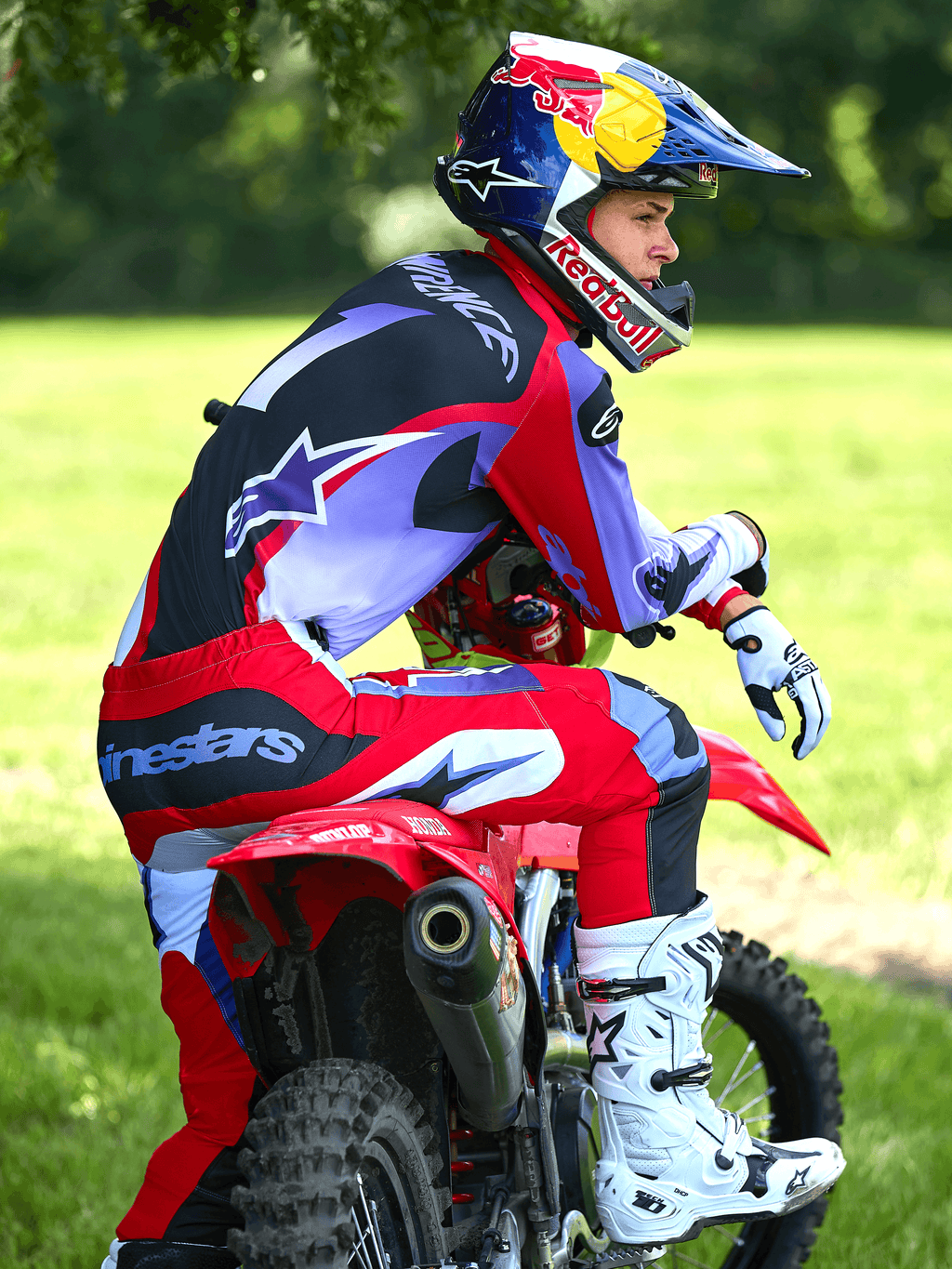 Piloto de motocross sentado numa mota de motocross vermelha, equipado com calças Alpinestars 2025 Fluid Wurx em roxo e vermelho vivo, camisola a condizer, botas Tech 10 brancas e pretas, luvas brancas e capacete Red Bull, conjunto completo de corrida todo-o-terreno num cenário ao ar livre