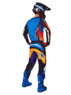 Piloto de motocross visto de costas a usar calças Alpinestars 2025 Fluid Wurx, design multicolor com detalhes em azul, laranja, amarelo e roxo, camisola a condizer, botas em azul vivo e capacete preto, equipamento completo de competição para todo-o-terreno