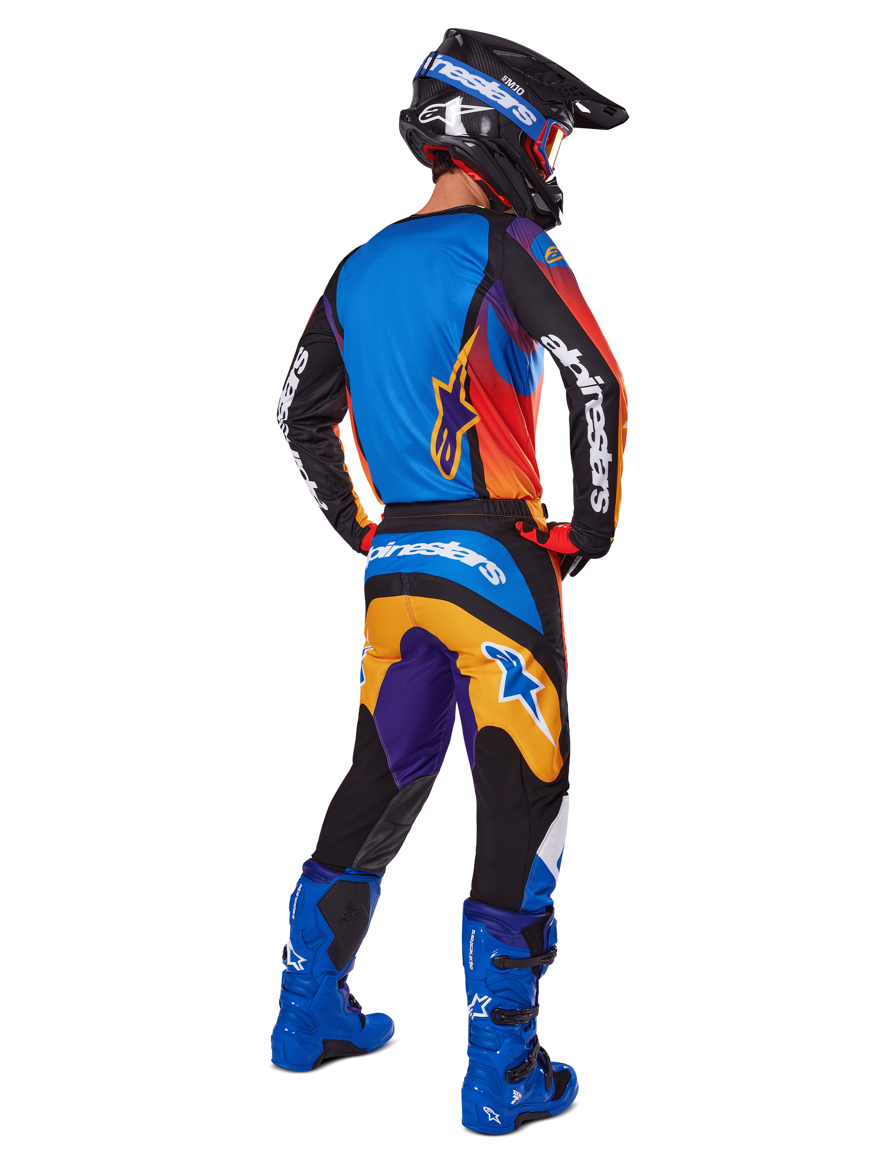 Piloto de motocross visto de costas a usar calças Alpinestars 2025 Fluid Wurx, design multicolor com detalhes em azul, laranja, amarelo e roxo, camisola a condizer, botas em azul vivo e capacete preto, equipamento completo de competição para todo-o-terreno