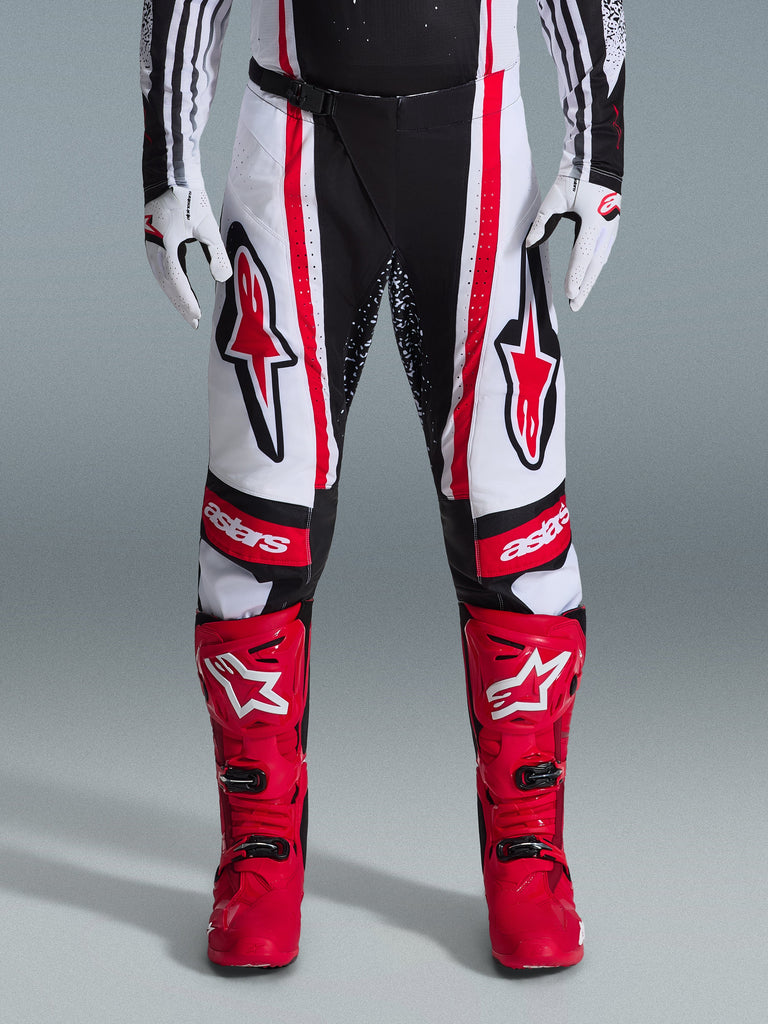 Alpinestars 2026 Techstar Nomur Pants, calças de motocross, pretas, brancas e vermelhas, usadas com botas vermelhas a condizer e luvas brancas, com logótipos vermelhos destacados nas coxas e marca 'astars' nos joelhos, design de corrida multi-painel para desempenho em todo-o-terreno