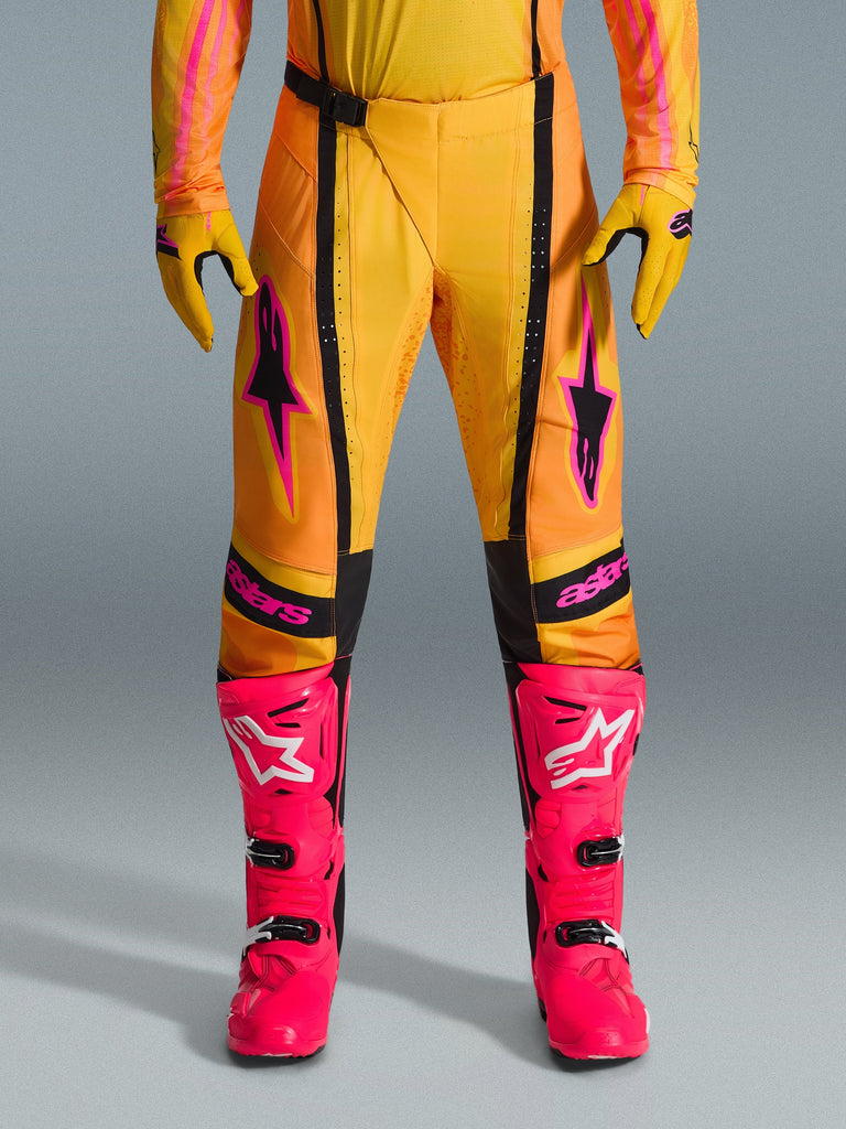 Alpinestars 2026 Techstar Nomur Pants, calças de motocross, laranja, amarelo e cor-de-rosa, usadas com luvas amarelas a condizer e botas de corrida cor-de-rosa vibrante, com painéis pretos e logótipo Alpinestars cor-de-rosa em destaque, equipamento técnico de corrida todo-o-terreno