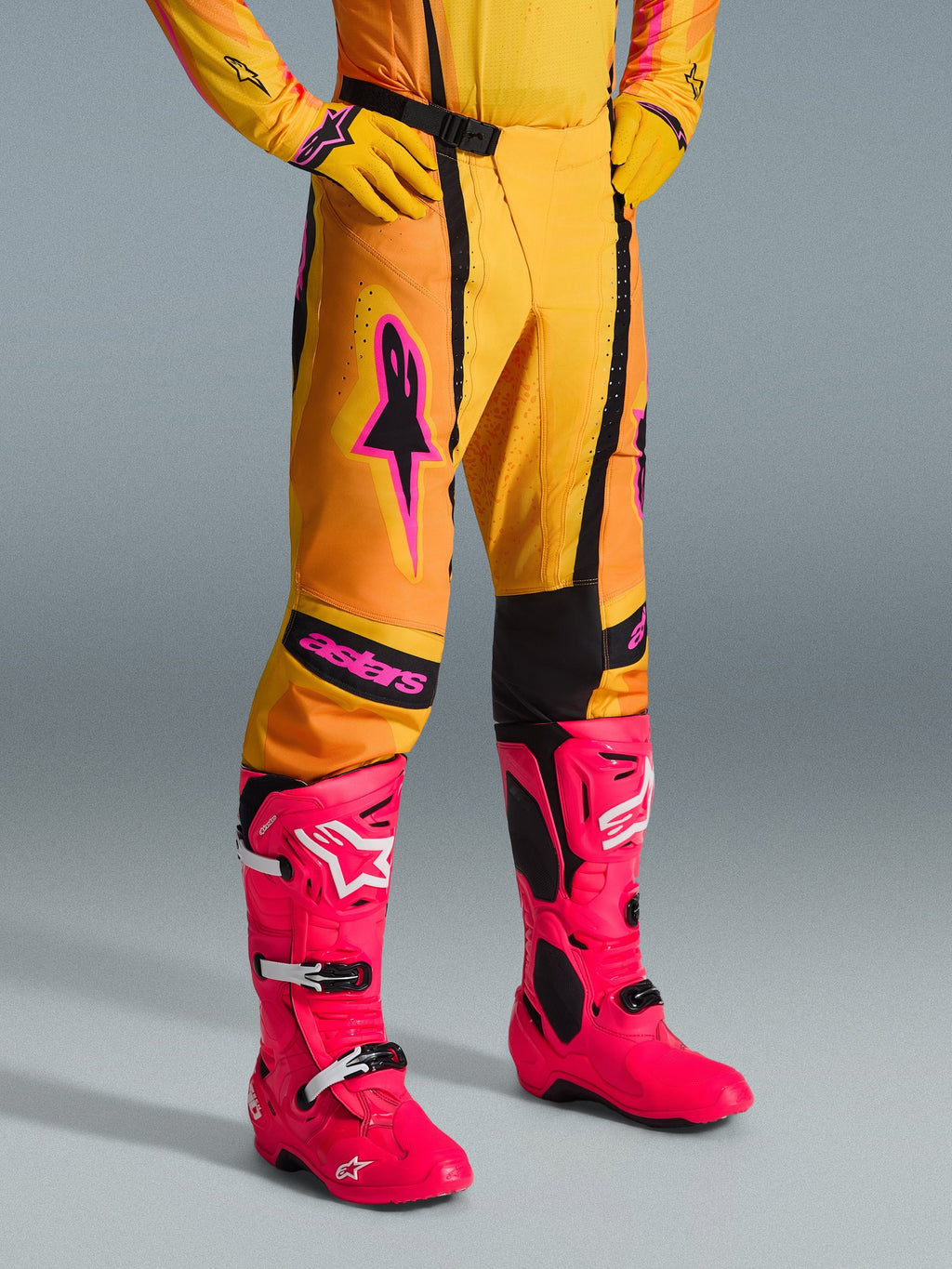 Alpinestars 2026 Techstar Nomur Pants, calças de motocross, laranja, amarelo e cor-de-rosa, usadas com camisola e luvas amarelas a condizer e botas cor-de-rosa vibrantes, corte atlético de corrida com logótipos cor-de-rosa e fivela magnética preta, equipamento vibrante para competição de todo-o-terreno