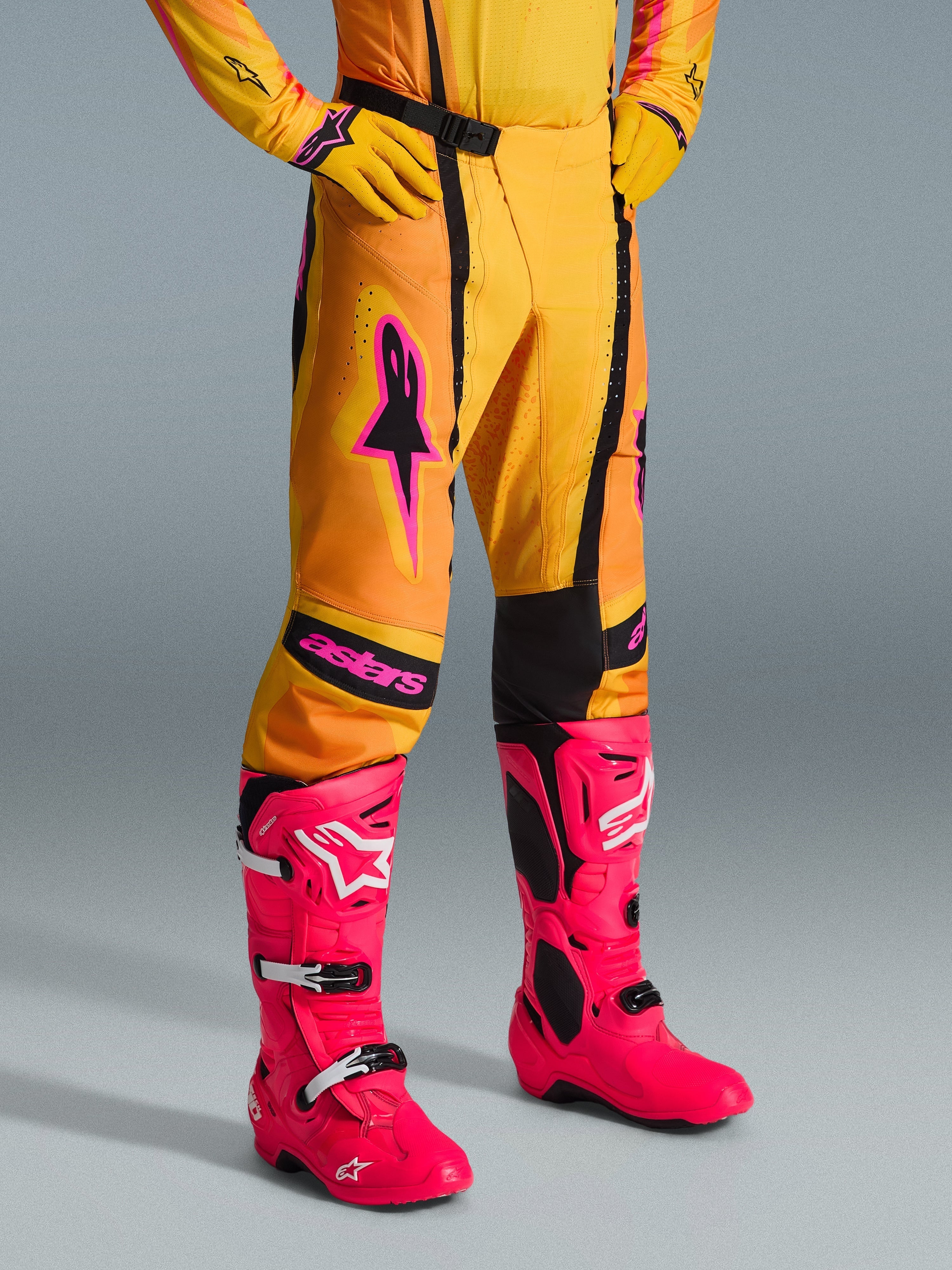 Alpinestars 2026 Techstar Nomur Pants, calças de motocross, laranja, amarelo e cor-de-rosa, usadas com camisola e luvas amarelas a condizer e botas cor-de-rosa vibrantes, corte atlético de corrida com logótipos cor-de-rosa e fivela magnética preta, equipamento vibrante para competição de todo-o-terreno