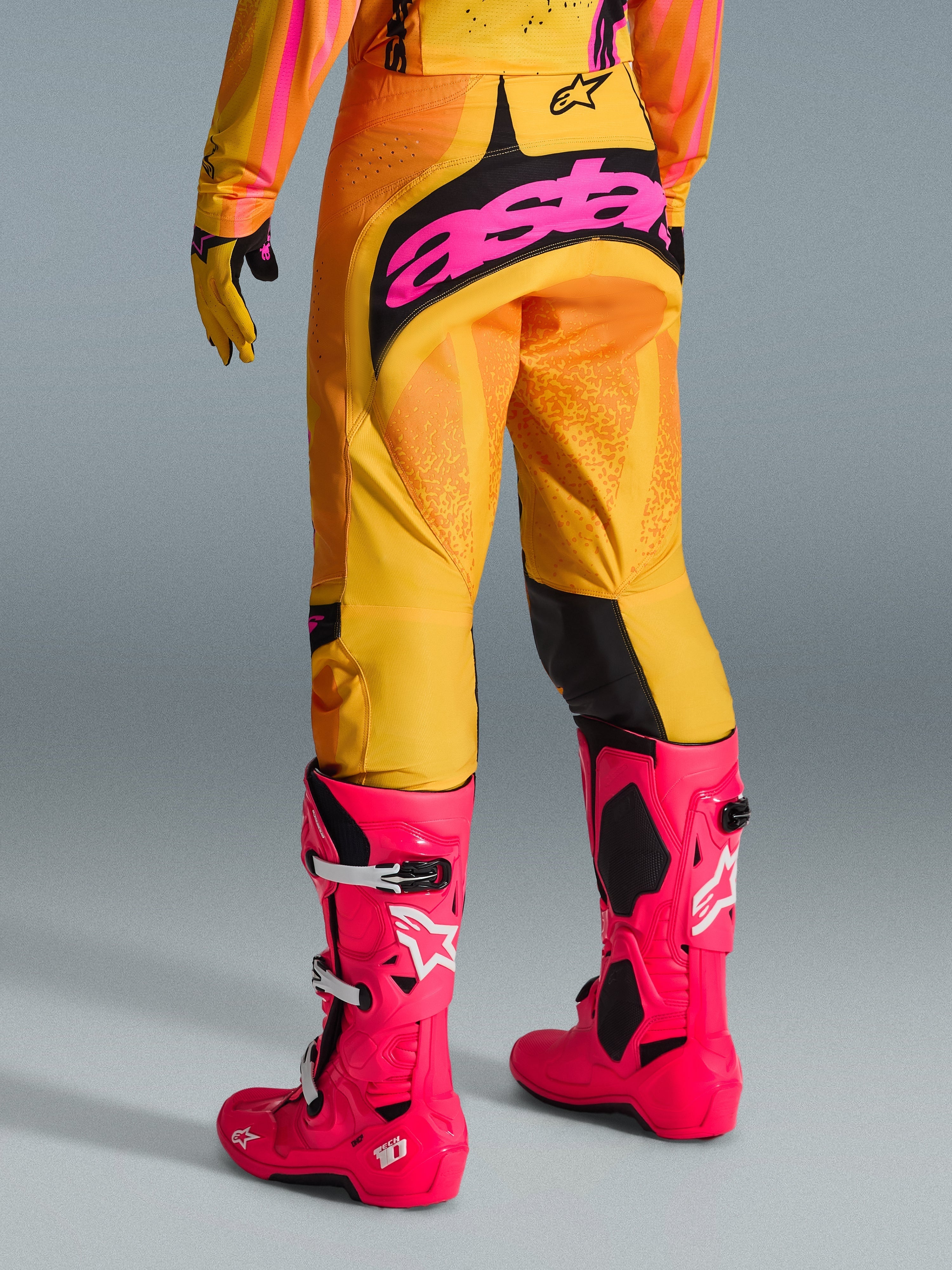 Alpinestars 2026 Techstar Nomur Pants, calças de motocross, laranja, amarelo e rosa, vista traseira, com logótipo 'astars' grande em rosa no assento, combinadas com botas Tech 10 cor-de-rosa vibrante e camisola a condizer, equipamento de corrida de alta performance para motocross