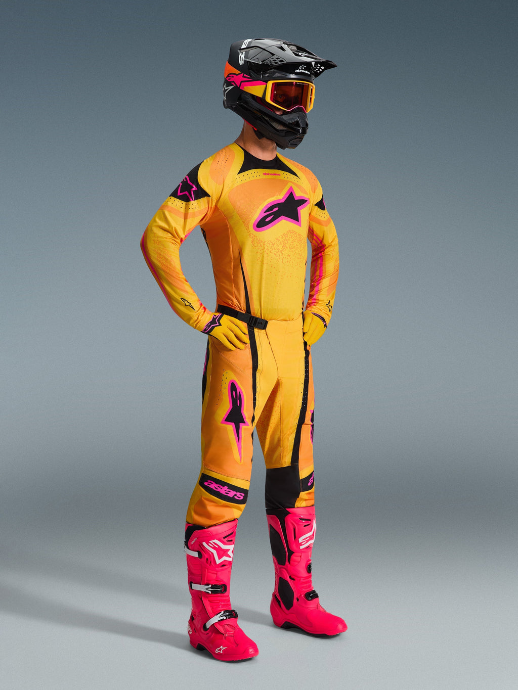 Piloto de motocross a usar calças Alpinestars 2026 Techstar Nomur em laranja, amarelo e rosa, conjunto completo de equipamento de corrida com camisola e luvas a condizer, botas cor-de-rosa vibrante, capacete preto com óculos cor de laranja, vista de corpo inteiro contra um fundo cinzento, equipamento profissional de corrida todo-o-terreno