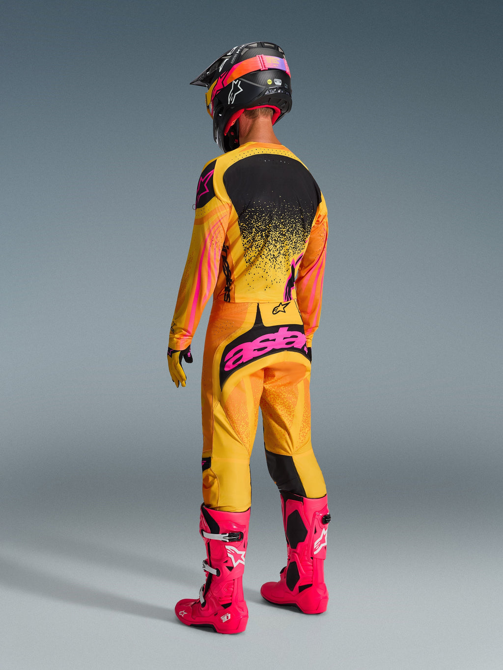 Piloto de motocross visto de costas com equipamento de corrida completo, Calças Techstar Nomur 2026 em Laranja Amarelo Rosa, marca 'astars' em rosa vibrante no assento, camisola a condizer, botas cor-de-rosa brilhante e capacete preto com detalhes em rosa, conjunto coordenado para competição off-road.