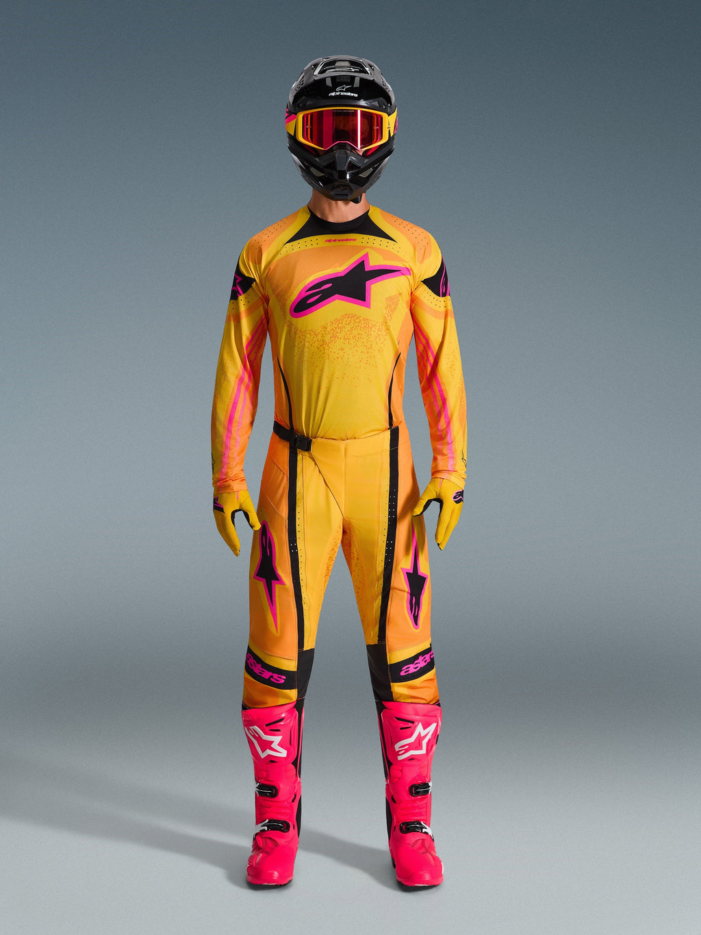 Piloto de motocross com o conjunto completo de equipamento Alpinestars 2026 Techstar Nomur, camisola e calças laranja, amarelo e cor-de-rosa, luvas amarelas e cor-de-rosa a condizer, botas de corrida cor-de-rosa vibrante, e capacete preto com óculos cor-de-rosa, conjunto de motocross coordenado de alta performance.