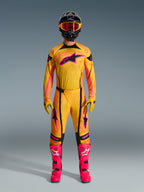 Piloto de motocross com o conjunto completo de equipamento Alpinestars 2026 Techstar Nomur, camisola e calças laranja, amarelo e cor-de-rosa, luvas amarelas e cor-de-rosa a condizer, botas de corrida cor-de-rosa vibrante, e capacete preto com óculos cor-de-rosa, conjunto de motocross coordenado de alta performance.