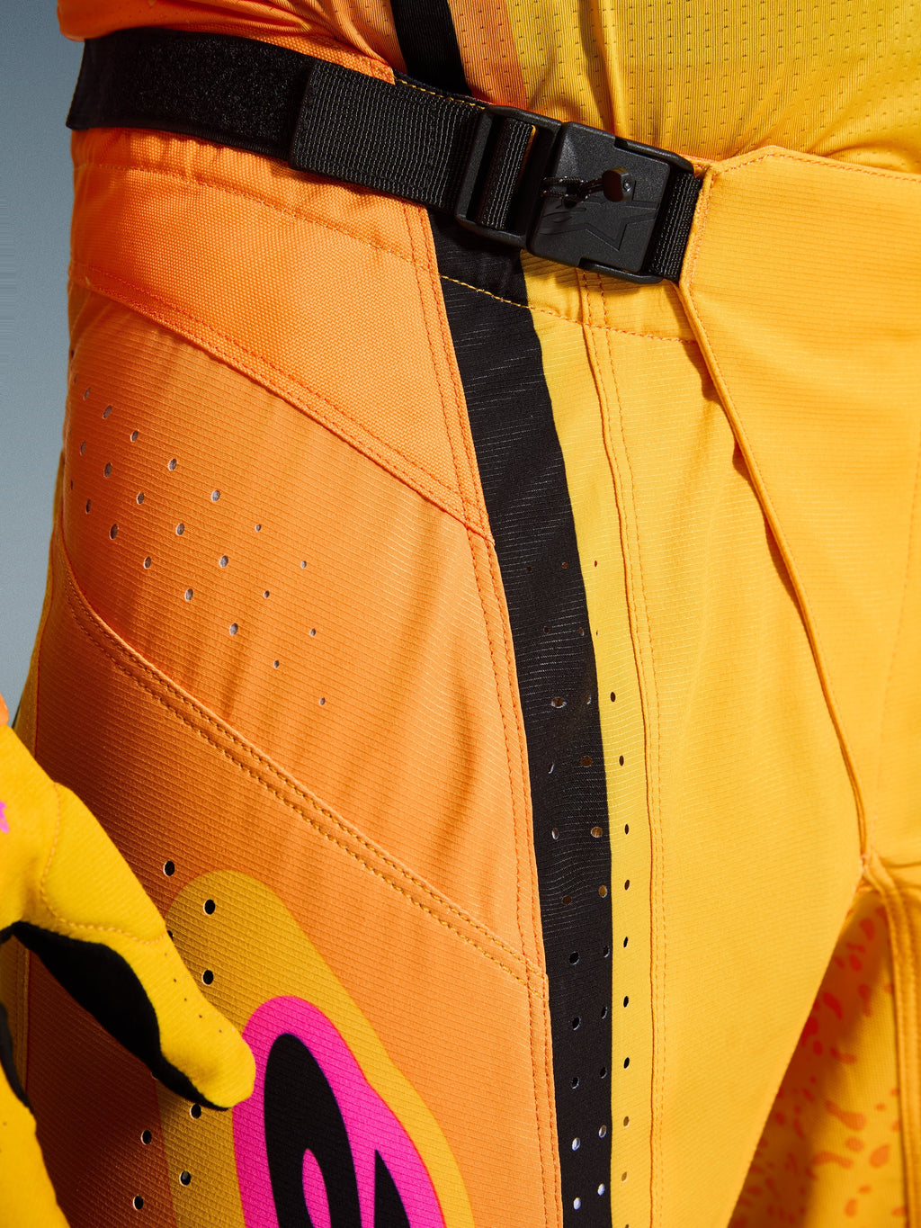 Alpinestars 2026 Techstar Nomur Pants, calças de motocross, cor de laranja, amarelo e rosa, pormenor da zona da cintura com o fecho de fivela magnética SX2, tira de ajuste preta e perfurações de ventilação cortadas a laser em material ripstop durável