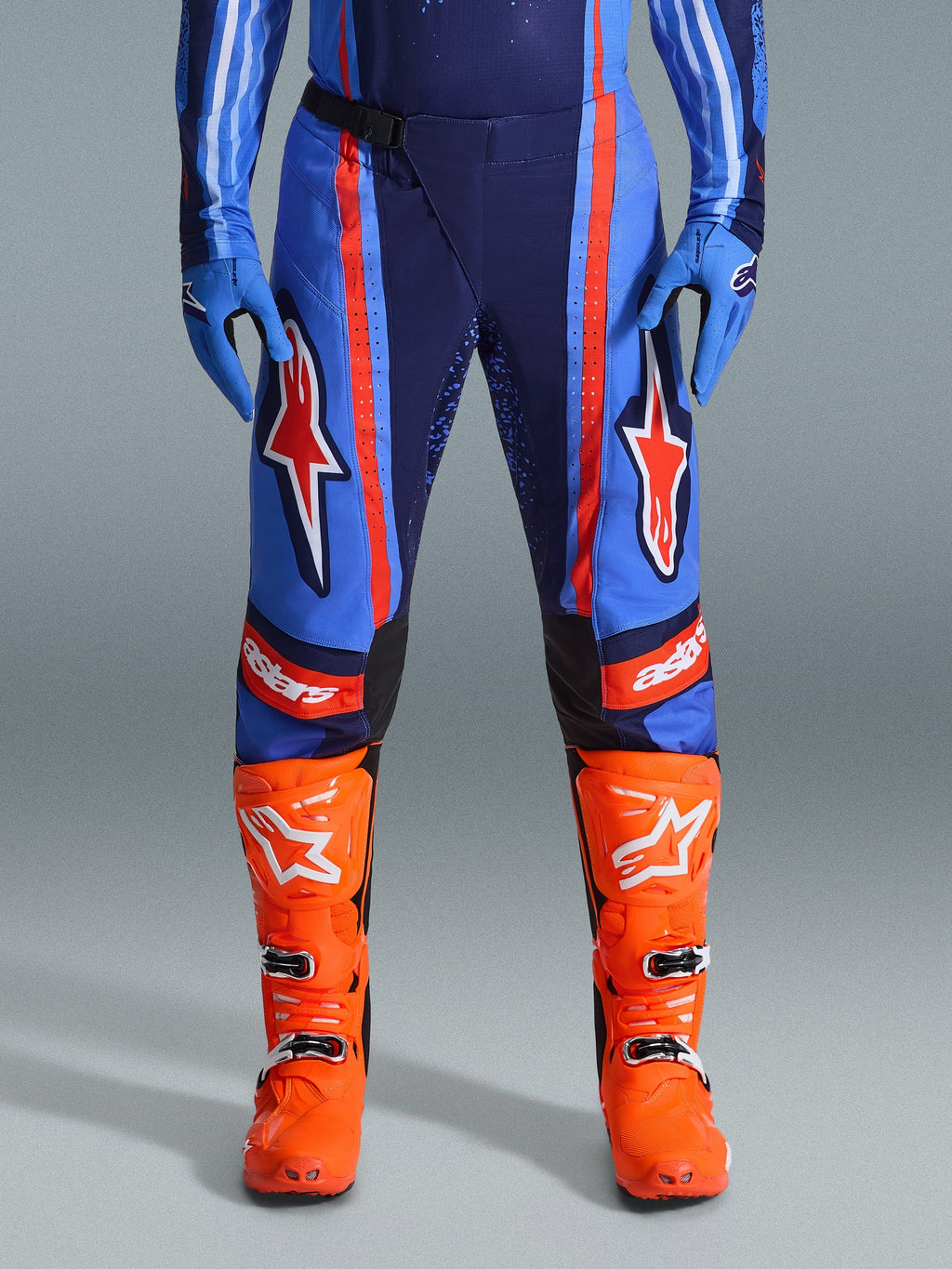 Alpinestars 2026 Techstar Nomur Pants, calças de motocross, azul-marinho escuro, laranja e azul UCLA, com riscas cor de laranja vibrantes e grandes logótipos de estrela brancos nas coxas, usadas com botas de competição cor de laranja brilhante e luvas azuis, corte atlético de corrida para performance off-road