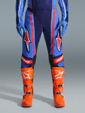 Alpinestars 2026 Techstar Nomur Pants, calças de motocross, azul-marinho escuro, laranja e azul UCLA, com riscas cor de laranja vibrantes e grandes logótipos de estrela brancos nas coxas, usadas com botas de competição cor de laranja brilhante e luvas azuis, corte atlético de corrida para performance off-road