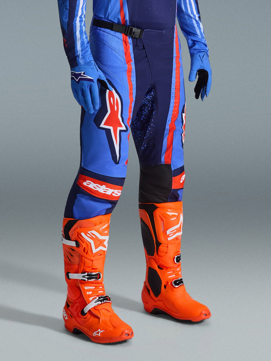 Alpinestars 2026 Techstar Nomur Pants, calças de motocross, azul-marinho, laranja e azul UCLA, usadas com botas de motocross cor de laranja vibrante e equipamento azul a condizer, com branding de destaque e construção multimaterial para desempenho off-road