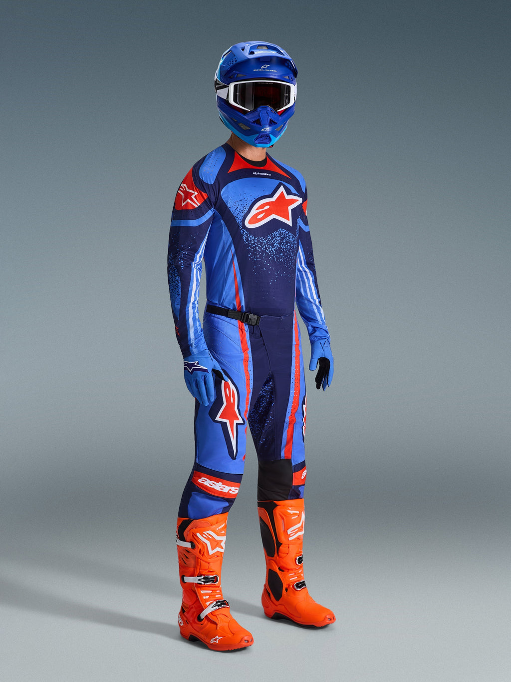 Alpinestars 2026 Techstar Nomur Pants, calças de motocross, azul-marinho escuro, laranja e azul UCLA, usadas por um piloto com equipamento de corrida completo, incluindo camisola a condizer, capacete azul e botas laranja vibrante, concebidas para performance em corridas off-road