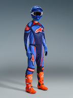 Alpinestars 2026 Techstar Nomur Pants, calças de motocross, azul-marinho escuro, laranja e azul UCLA, usadas por um piloto com equipamento de corrida completo, incluindo camisola a condizer, capacete azul e botas laranja vibrante, concebidas para performance em corridas off-road