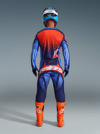 Piloto de motocross visto de costas a usar as calças Alpinestars 2026 Techstar Nomur em azul marinho escuro, laranja e azul UCLA, combinadas com uma camisola a condizer e botas cor de laranja vibrante, conjunto de equipamento profissional de off-road.