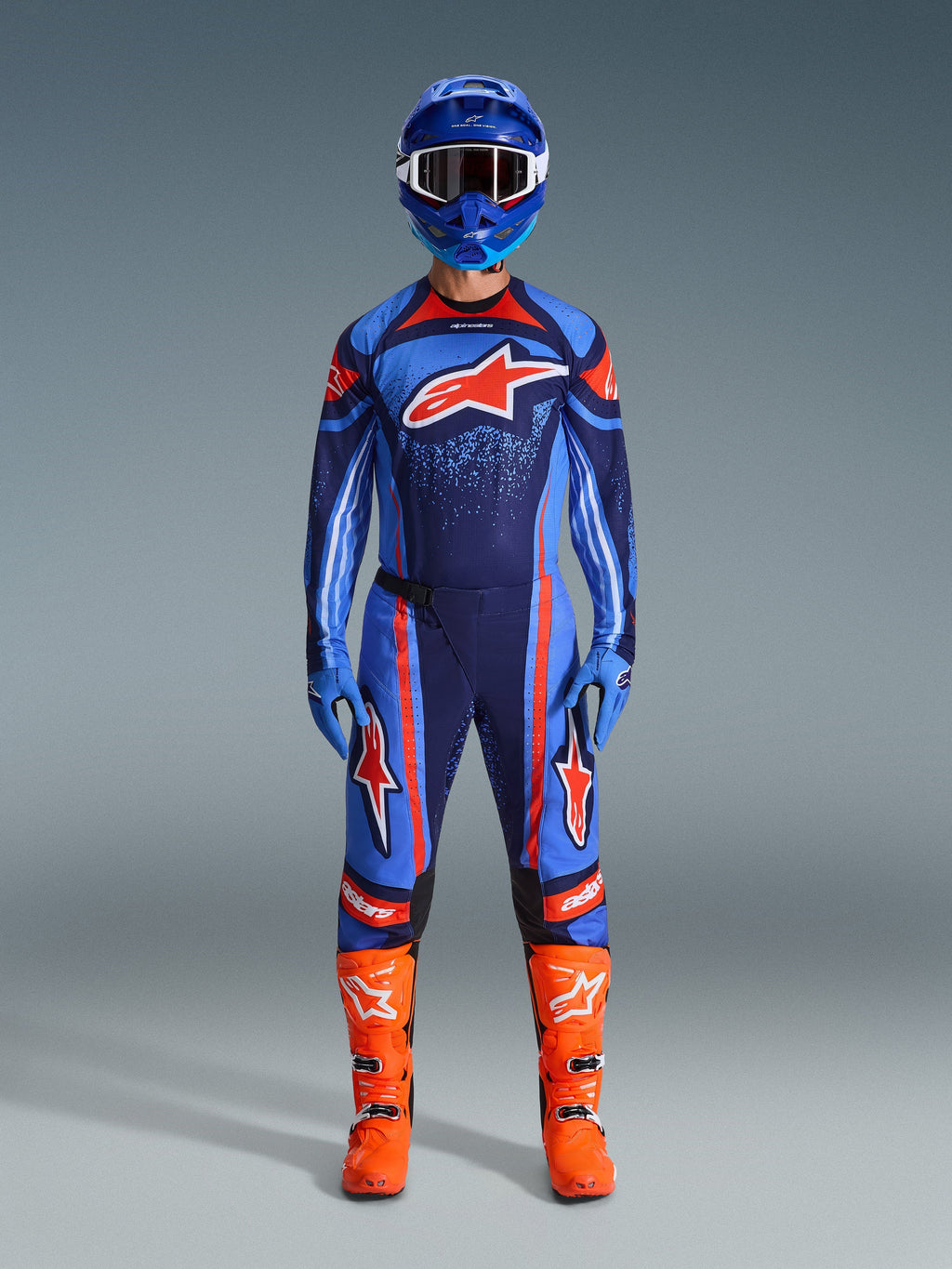 Conjunto de equipamento Alpinestars 2026 Techstar Nomur, piloto de motocross com kit de corrida completo, azul-escuro com detalhes em laranja e azul UCLA, camisola e calças a condizer com logótipos grandes, capacete azul e botas laranja vibrante, equipamento de competição todo-o-terreno coordenado.