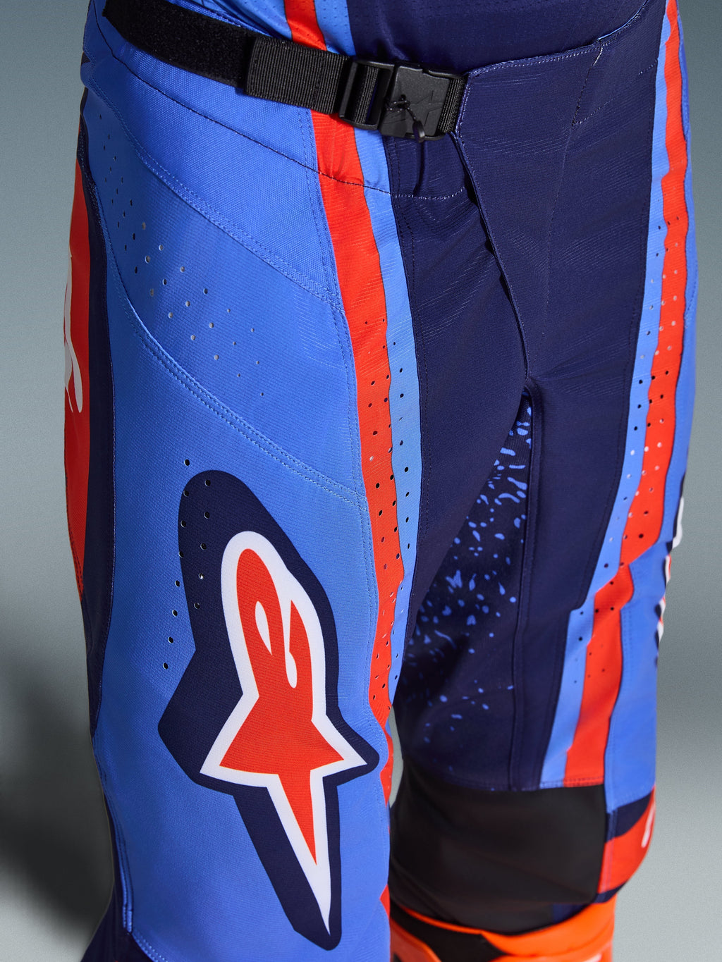 Alpinestars 2026 Techstar Nomur Pants, calças de motocross em azul marinho escuro, laranja e azul UCLA, plano aproximado que mostra o fecho de cintura com fivela magnética SX2, perfurações a laser na coxa e o icónico logótipo de estrela, equipamento de corrida off-road durável.