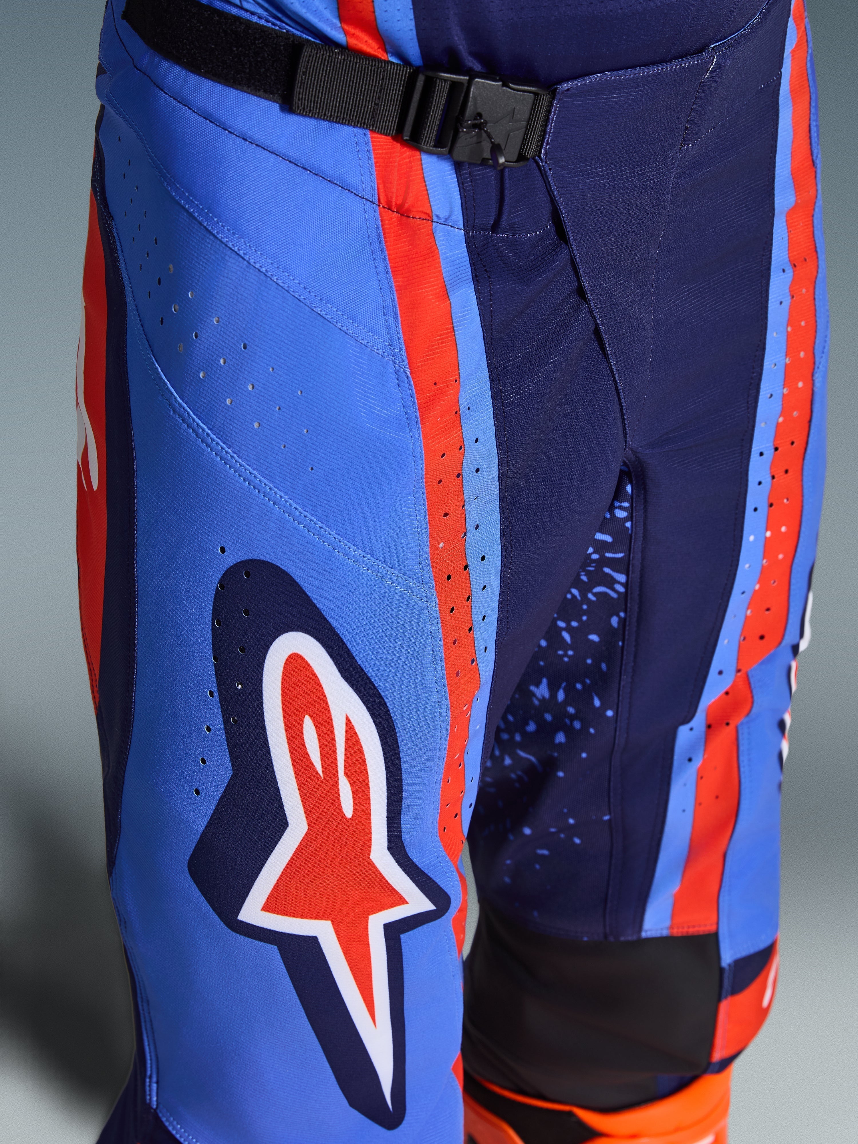 Alpinestars 2026 Techstar Nomur Pants, calças de motocross em azul marinho escuro, laranja e azul UCLA, plano aproximado que mostra o fecho de cintura com fivela magnética SX2, perfurações a laser na coxa e o icónico logótipo de estrela, equipamento de corrida off-road durável.
