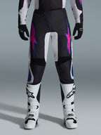 Alpinestars 2026 Fluid Grid Pants, calças de motocross, pretas com desenhos de estrelas em roxo e azul, usadas com botas de motocross brancas e luvas pretas, com painéis reforçados e um ajuste desportivo para todo-o-terreno, fundo cinzento