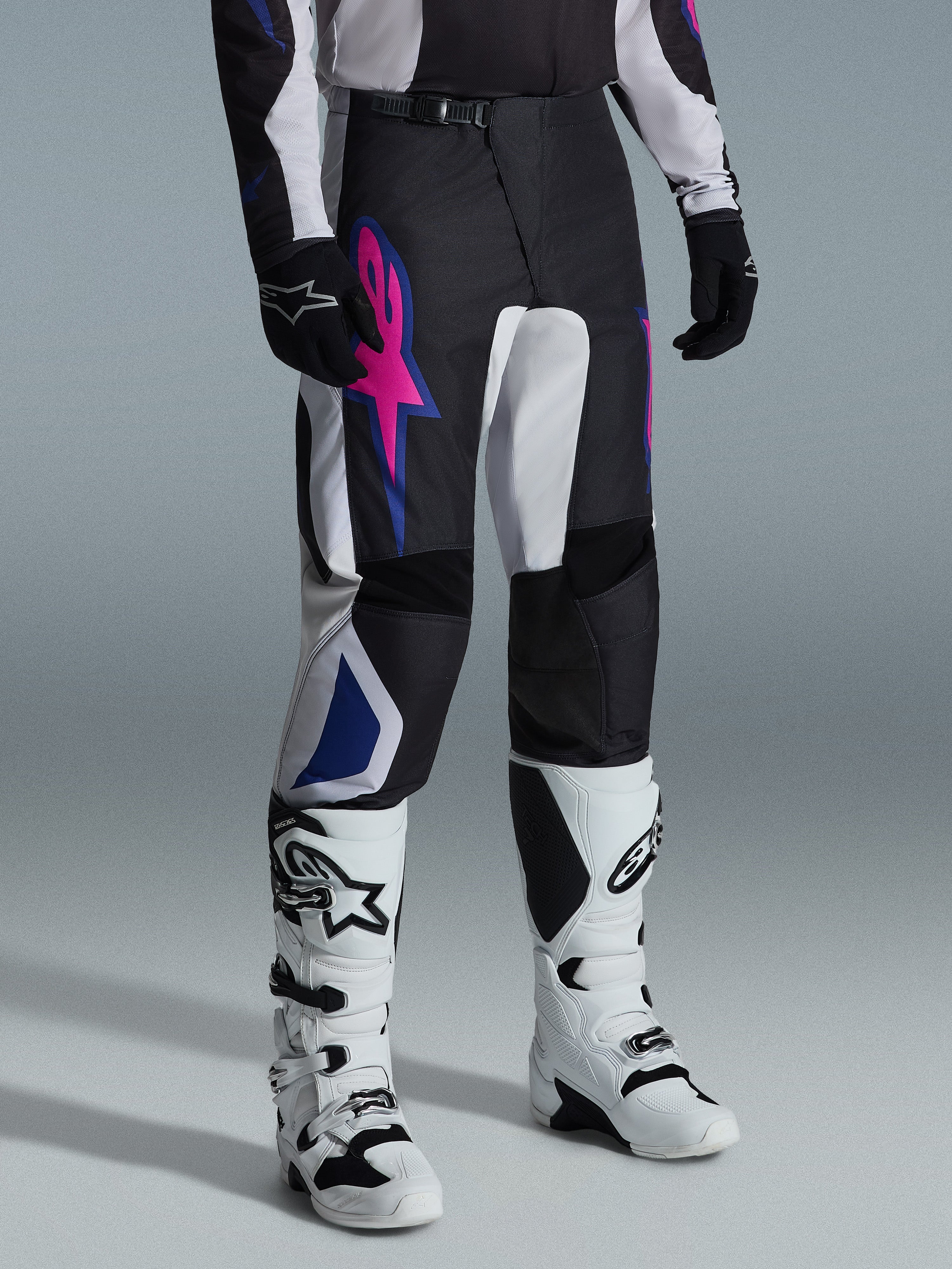 Alpinestars 2026 Fluid Grid Pants, calças de motocross, em preto, cinzento claro e roxo, com um logótipo de estrela rosa vibrante e detalhes geométricos azuis, usadas com botas de motocross brancas e luvas pretas, equipamento ergonómico para condução todo-o-terreno