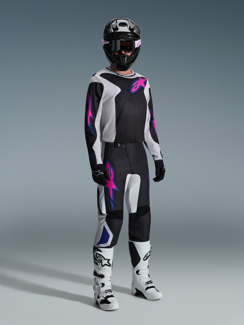 Piloto de motocross com equipamento de corrida completo, calças Alpinestars 2026 Fluid Grid em preto, cinzento claro e roxo, camisola a condizer com gráficos cor-de-rosa e azuis, capacete e óculos pretos, botas brancas e pretas, e luvas pretas, conjunto coordenado para corridas todo-o-terreno