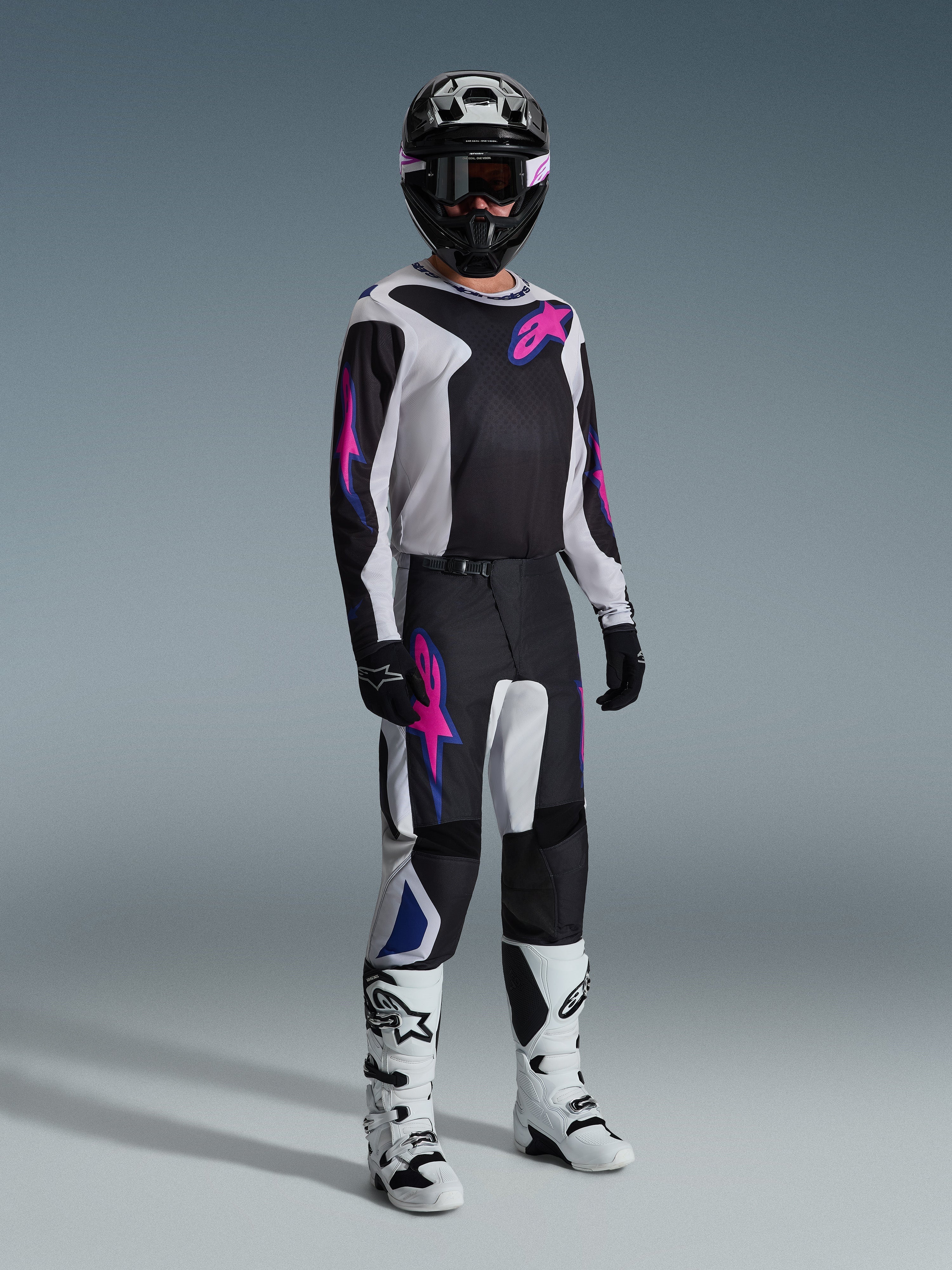 Piloto de motocross com equipamento de corrida completo, calças Alpinestars 2026 Fluid Grid em preto, cinzento claro e roxo, camisola a condizer com gráficos cor-de-rosa e azuis, capacete e óculos pretos, botas brancas e pretas, e luvas pretas, conjunto coordenado para corridas todo-o-terreno