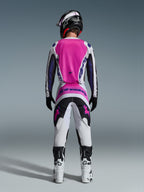 Piloto de motocross visto de costas a usar calças Alpinestars 2026 Fluid Grid e camisola a condizer, rosa, branco e preto com logótipos em roxo, capacete preto, botas brancas e pretas, equipamento de corrida coordenado para motocross todo-o-terreno