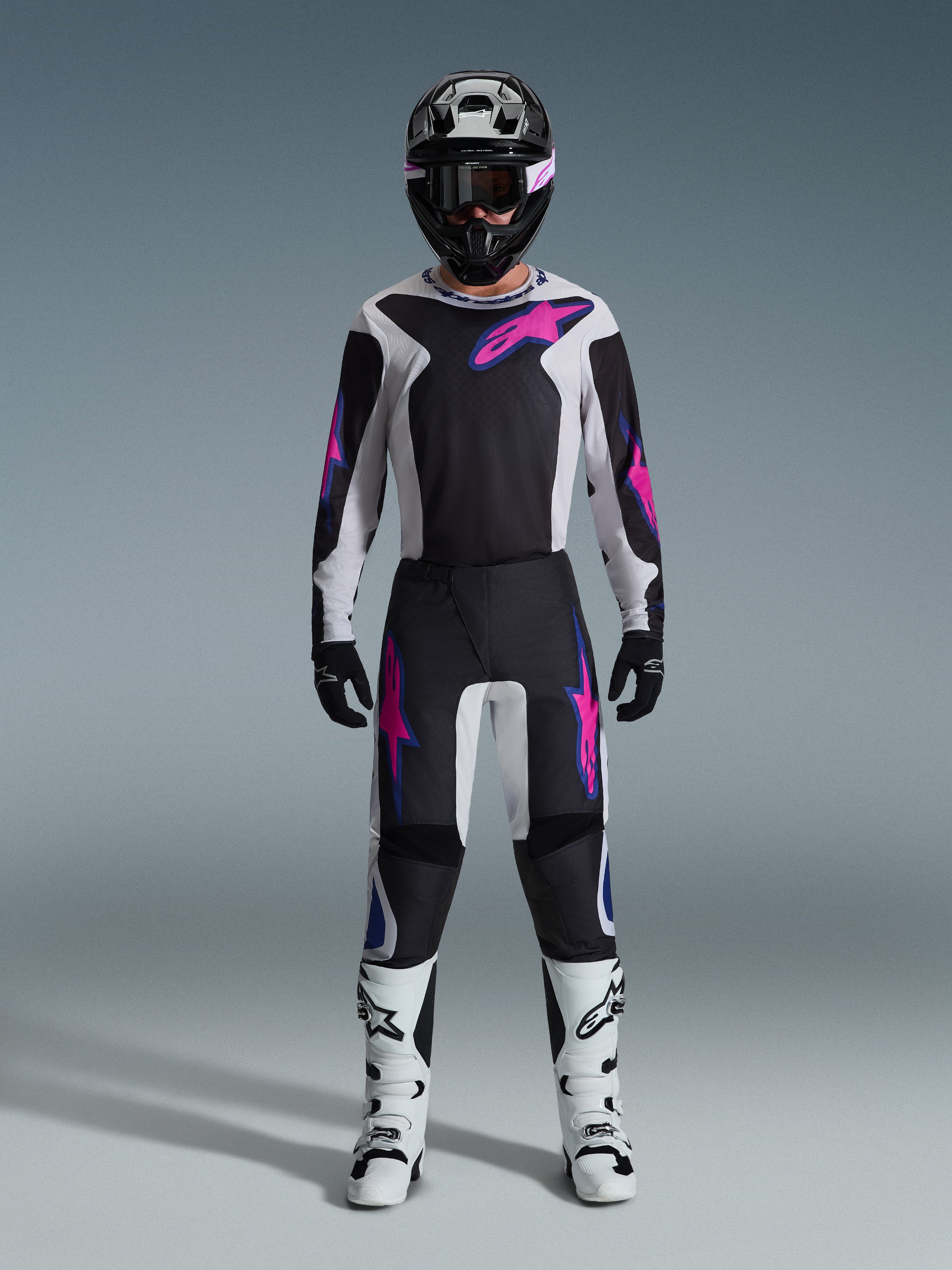 Piloto de motocross a usar o Conjunto de Equipamento Alpinestars 2026 Fluid Grid, camisola preta e cinzenta clara com logótipos cor-de-rosa vibrantes, calças pretas e brancas com grafismos cor-de-rosa e azuis a combinar, capacete preto com óculos cor-de-rosa, botas brancas e pretas, conjunto completo de equipamento de corrida todo-o-terreno