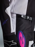 Alpinestars 2026 Fluid Grid Pants, calças de motocross, em preto, cinzento claro e roxo, grande plano apresentando o sistema de fecho de catraca na cintura e um grafismo arrojado em cor-de-rosa e roxo na anca, usadas com uma camisola a condizer e luva preta