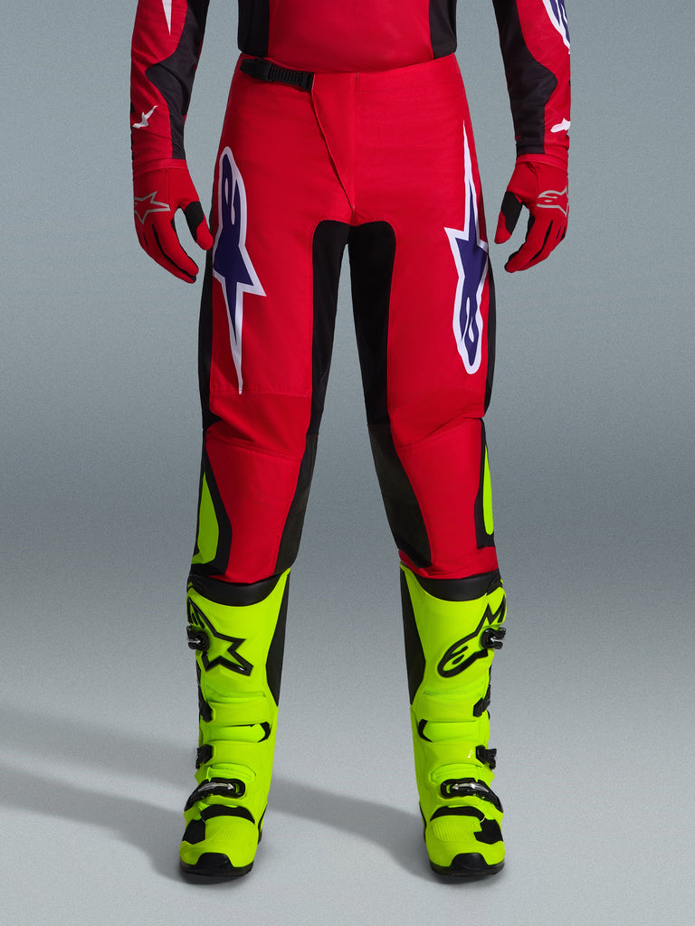Alpinestars 2026 Fluid Grid Pants, calças de motocross, vermelhas com painéis pretos e detalhes em amarelo néon, com grandes logótipos de estrela em branco e roxo nas coxas, usadas com luvas vermelhas e botas amarelo néon, construção durável para competição todo-o-terreno