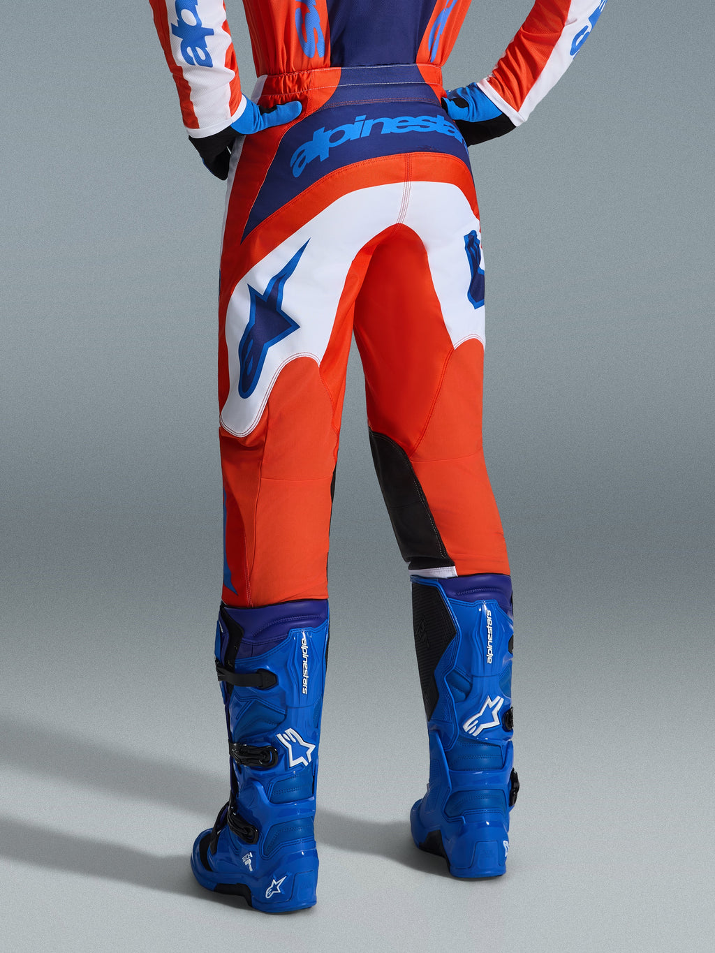 Alpinestars 2026 Fluid Grid Pants, calças de motocross, laranja e azul UCLA com painéis brancos, vista traseira mostrando a marca azul em destaque no assento e o logótipo de estrela, combinadas com botas de motocross azuis a condizer, equipamento de corrida off-road profissional