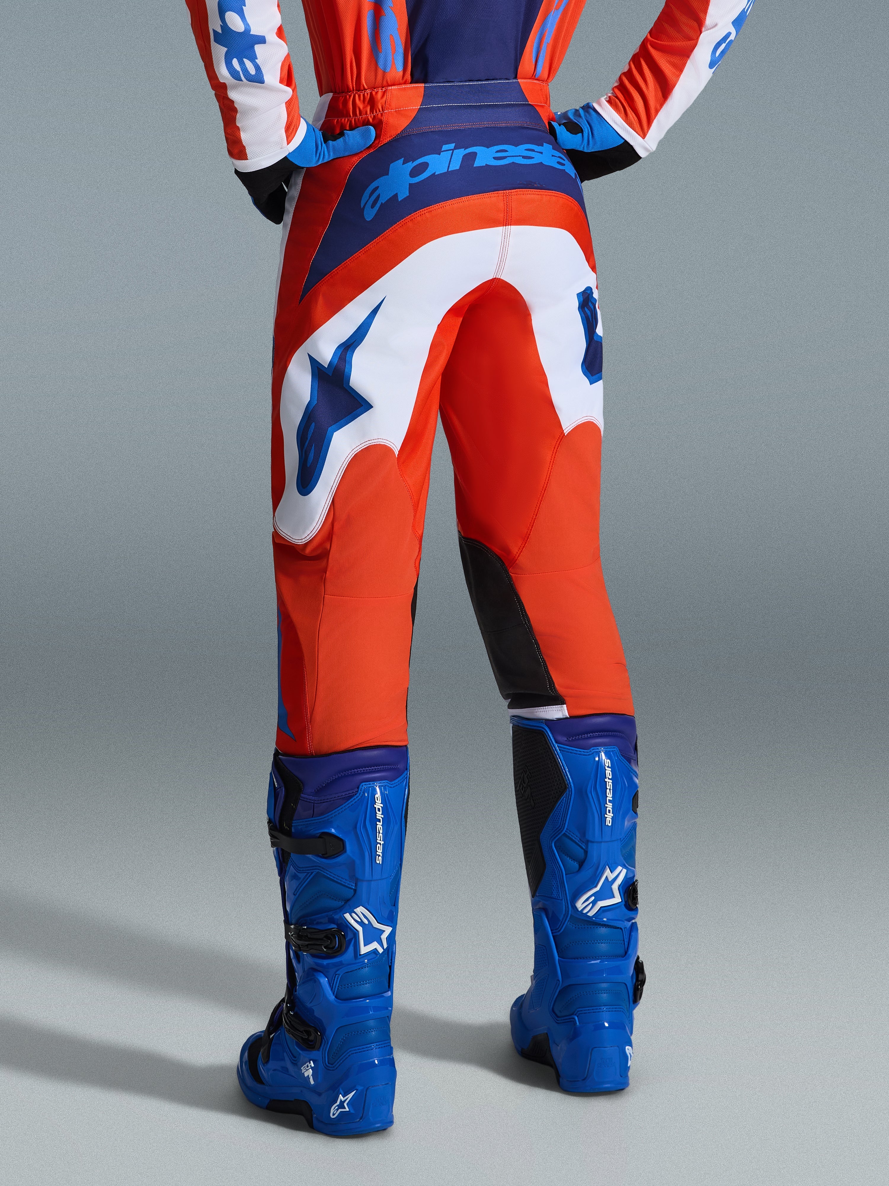 Alpinestars 2026 Fluid Grid Pants, calças de motocross, laranja e azul UCLA com painéis brancos, vista traseira mostrando a marca azul em destaque no assento e o logótipo de estrela, combinadas com botas de motocross azuis a condizer, equipamento de corrida off-road profissional