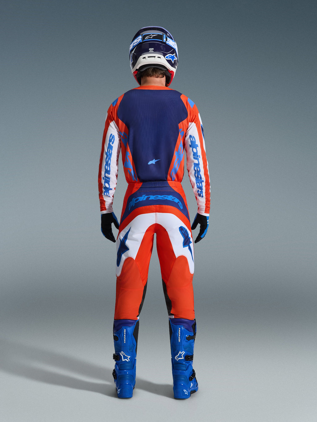 Alpinestars 2026 Fluid Grid Pants, calças de motocross, cor de laranja e azul UCLA, vista traseira completa de um piloto a usar um conjunto de equipamento de corrida completo com camisola a condizer, botas azuis e capacete, com branding proeminente e painéis ergonómicos para condução off-road.