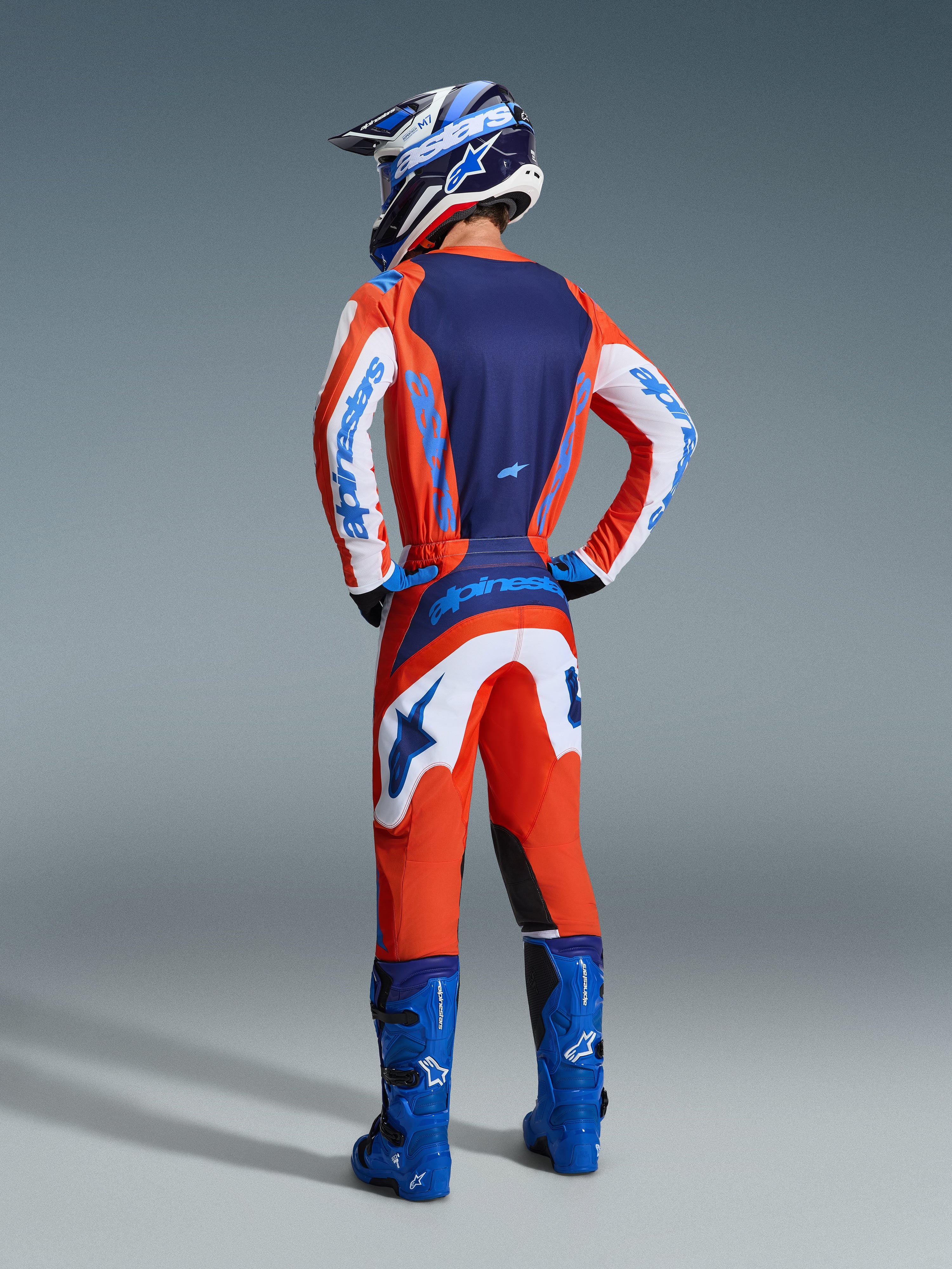 Piloto de motocross visto de costas a usar as calças Alpinestars 2026 Fluid Grid em laranja e azul UCLA, combinadas com uma camisola a condizer, botas de competição azuis e capacete azul e branco, conjunto completo de equipamento para condução off-road.