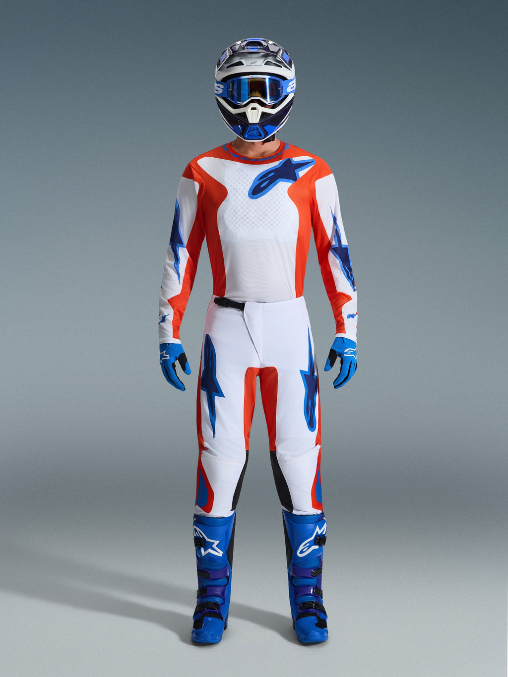Piloto de motocross com equipamento de corrida completo, calças Alpinestars 2026 Fluid Grid em Laranja e UCLA Blue, camisola branca com mangas laranja e logótipos azuis, capacete e óculos azuis a condizer, botas e luvas azuis, conjunto coordenado para corridas off-road