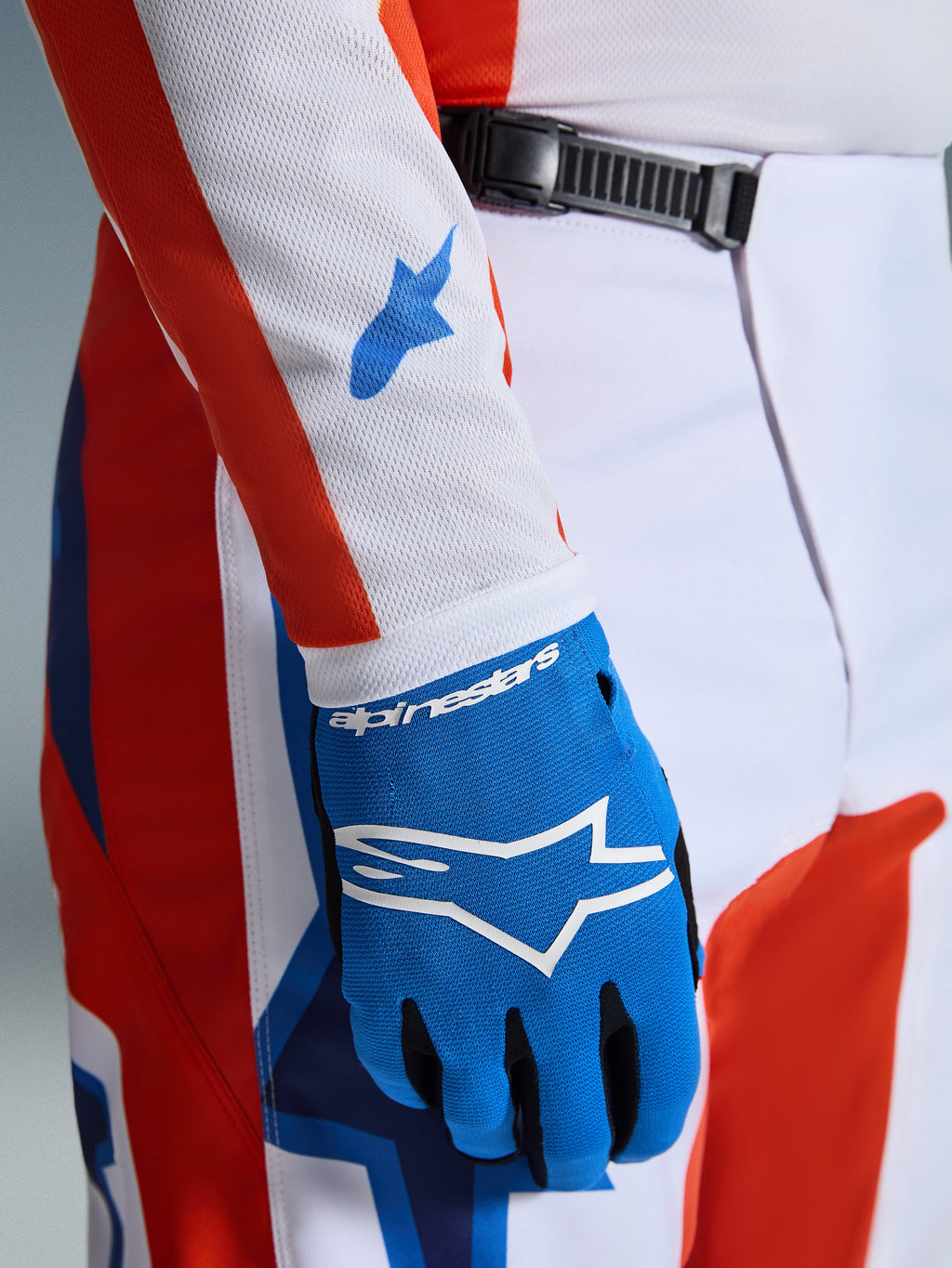 Alpinestars 2026 Fluid Grid Pants, calças de motocross, laranja e azul UCLA, grande plano com uma luva azul a condizer e manga de camisola em malha branca com logótipos azuis, destacando o fecho de catraca na cintura e o ajuste atlético