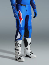 Alpinestars 2026 Fluid Grid Pants, calças de motocross, azul e vermelho, usadas com botas de motocross brancas e luvas azuis, com um grande logótipo de estrela vermelha na coxa e painéis pretos no interior do joelho, concebidas para condução off-road
