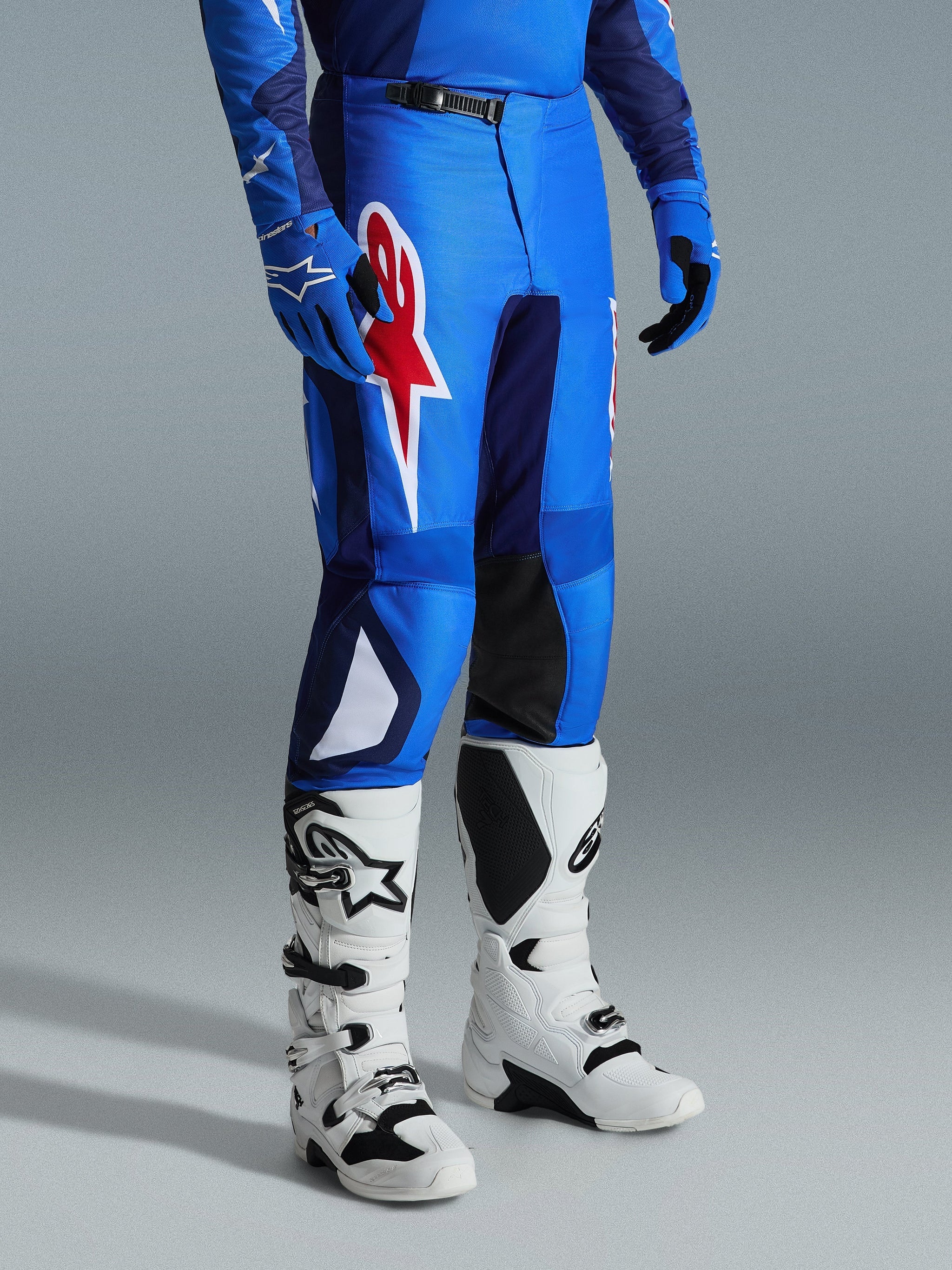 Alpinestars 2026 Fluid Grid Pants, calças de motocross, azul e vermelho, usadas com botas de motocross brancas e luvas azuis, com um grande logótipo de estrela vermelha na coxa e painéis pretos no interior do joelho, concebidas para condução off-road