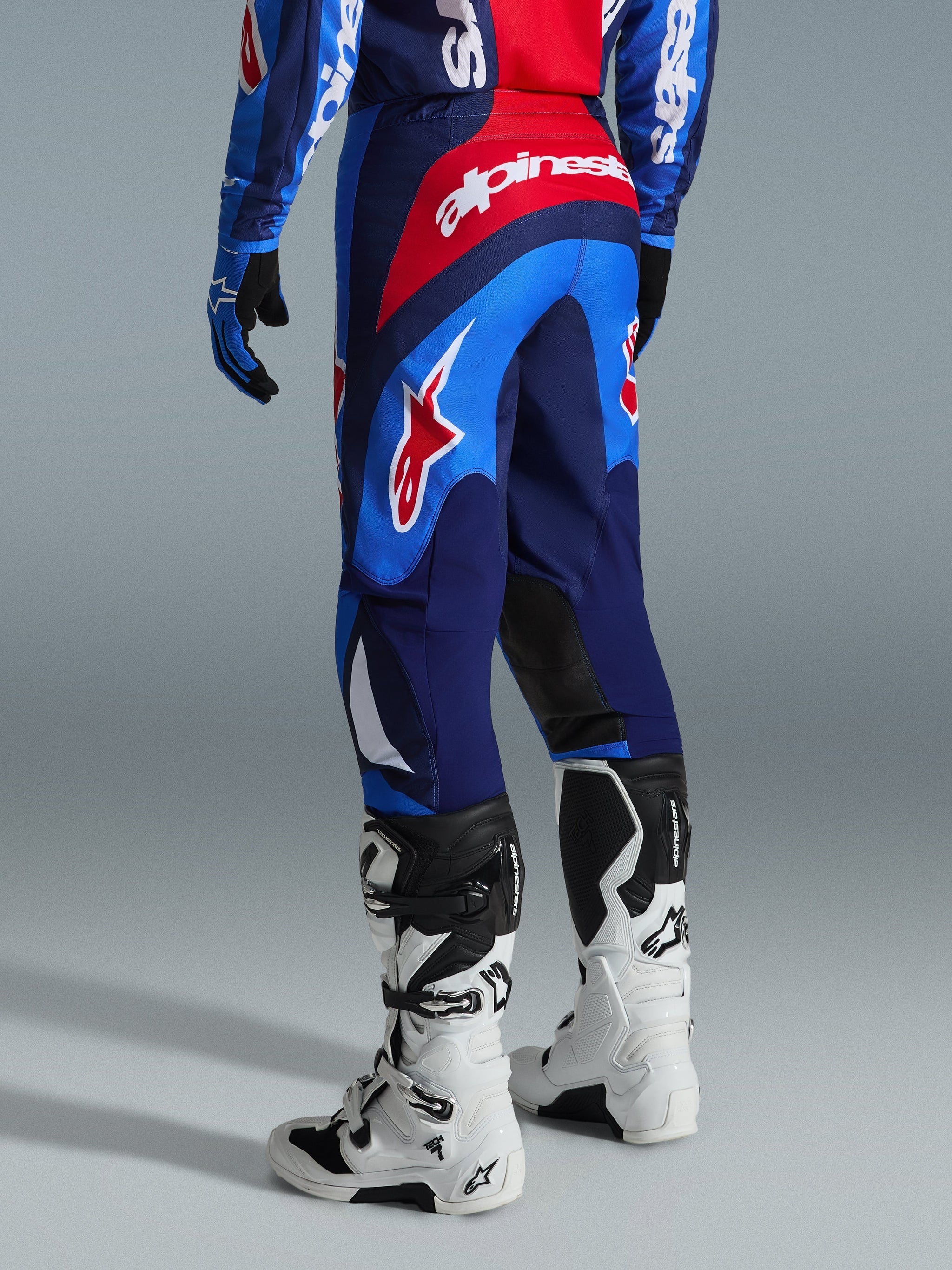 Alpinestars 2026 Fluid Grid Pants, calças de motocross, azuis e vermelhas, vista traseira mostrando a marca Alpinestars em destaque e design ergonómico, usadas com botas Tech 7 brancas e pretas e camisola a condizer, equipamento de alto desempenho para condução todo-o-terreno