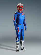 Piloto de motocross a usar equipamento de corrida completo, camisola azul e vermelha com padrão de grelha e logótipo Alpinestars, calças Fluid Grid 2026 em azul e vermelho com painéis internos pretos nos joelhos, capacete vermelho e branco com óculos, botas brancas e pretas, luvas azuis, conjunto coordenado para corridas off-road
