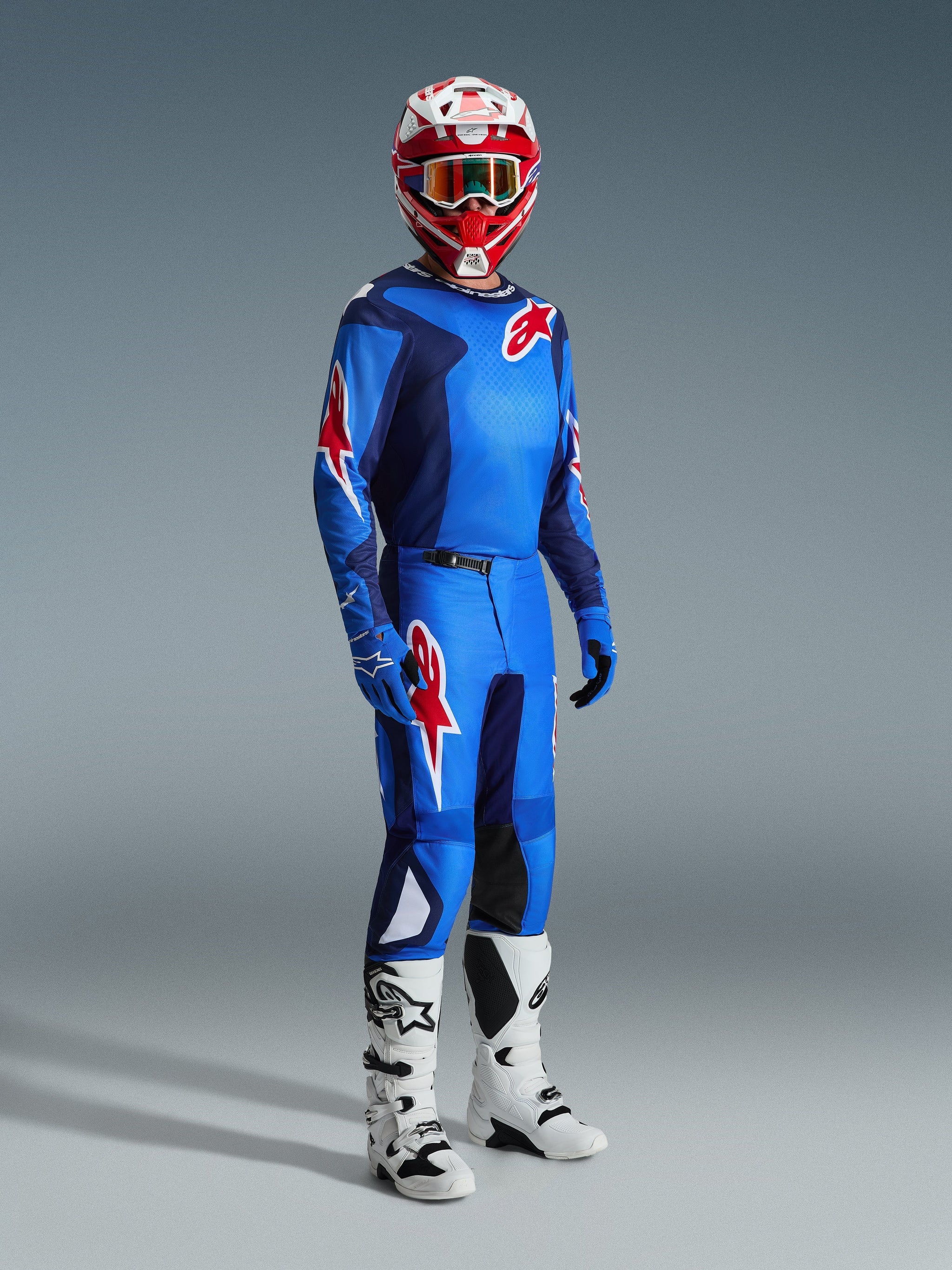 Piloto de motocross a usar equipamento de corrida completo, camisola azul e vermelha com padrão de grelha e logótipo Alpinestars, calças Fluid Grid 2026 em azul e vermelho com painéis internos pretos nos joelhos, capacete vermelho e branco com óculos, botas brancas e pretas, luvas azuis, conjunto coordenado para corridas off-road