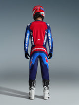 Piloto de motocross visto de costas com equipamento de corrida completo, Calças Fluid Grid 2026 em Azul e Vermelho, camisola vermelha e azul a condizer com logótipo Alpinestars, capacete vermelho e azul, botas brancas e pretas, conjunto coordenado para corridas off-road.