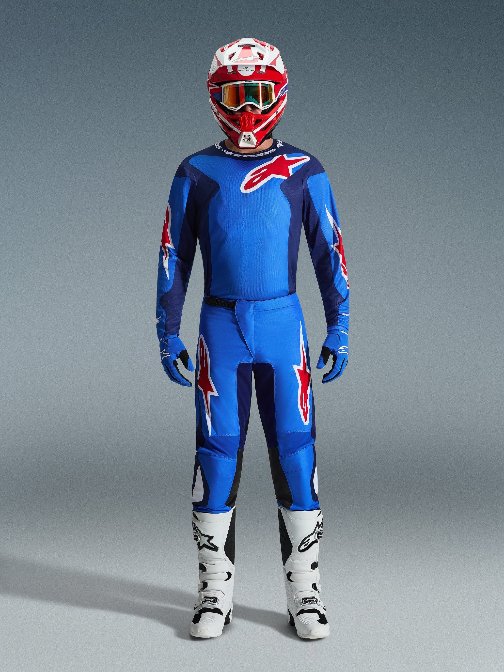 Piloto de motocross a usar calças Alpinestars 2026 Fluid Grid em azul e vermelho, camisola azul a condizer com logótipo Astars vermelho, capacete vermelho e branco com óculos, luvas azuis e botas de motocross brancas, equipamento completo e coordenado de corrida off-road.