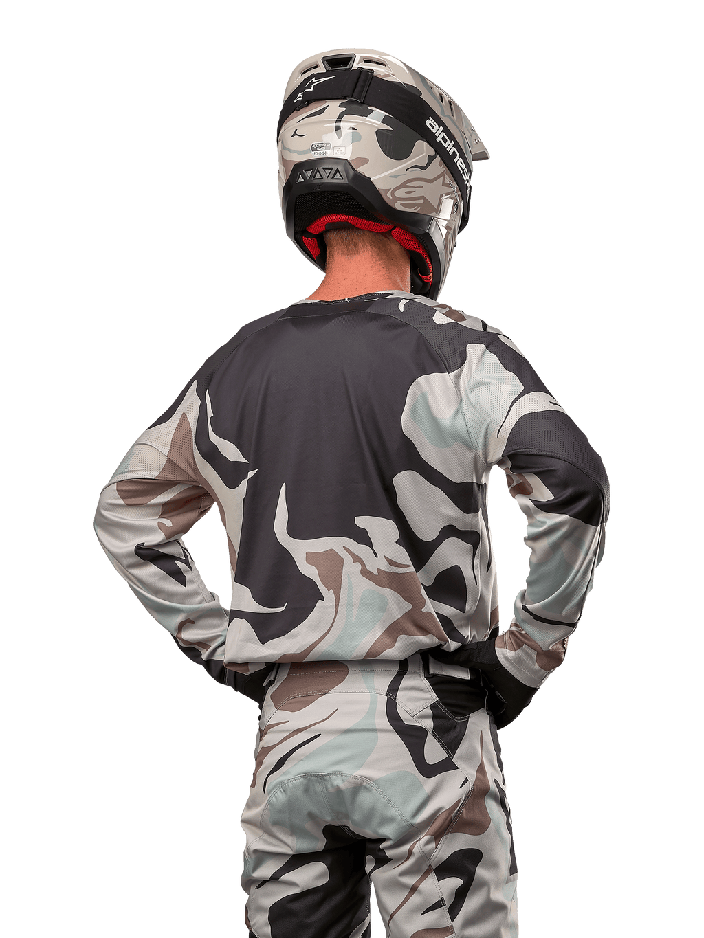 Piloto de motocross visto de costas a usar a camisola Alpinestars 2024 Racer Tactical em Iron Camo Dust Gray, calças e capacete de camuflagem a condizer, mãos nas ancas, equipamento de corrida off-road coordenado.