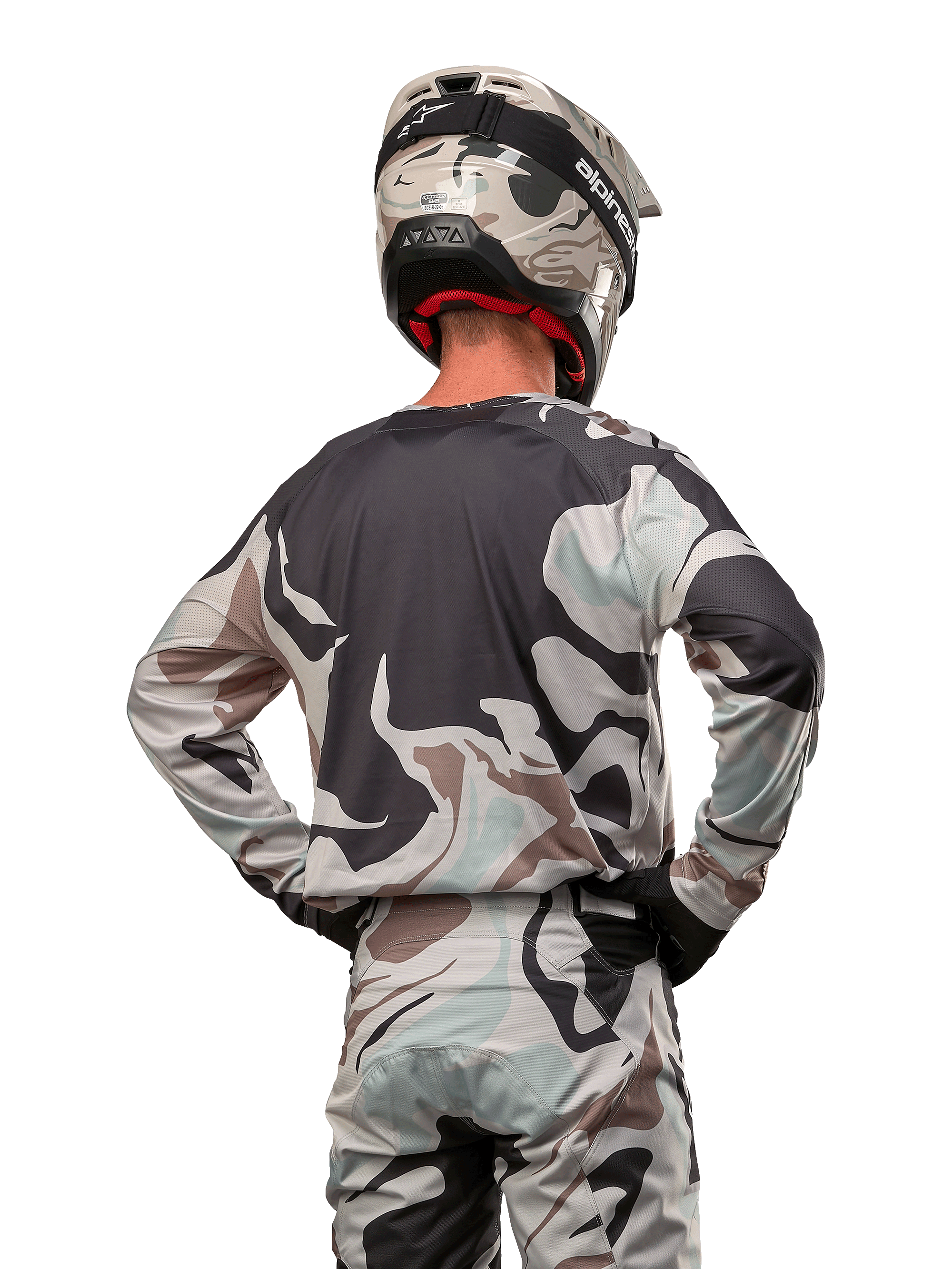 Piloto de motocross visto de costas a usar a camisola Alpinestars 2024 Racer Tactical em Iron Camo Dust Gray, calças e capacete de camuflagem a condizer, mãos nas ancas, equipamento de corrida off-road coordenado.