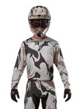 Alpinestars 2024 Racer Tactical Jersey, camisola de motocross, padrão camuflado Iron Camo Dust Gray, piloto a usar capacete e óculos a condizer com luvas pretas, equipamento técnico para corridas fora de estrada
