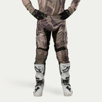 Alpinestars 2024 Racer Tactical Pants, calças de motocross, camuflado verde militar e castanho, com grandes logótipos Alpinestars brancos nas coxas e painéis pretos no interior das pernas, usadas com botas de motocross brancas e pretas, luvas pretas e camisola camuflada a condizer, vista frontal.