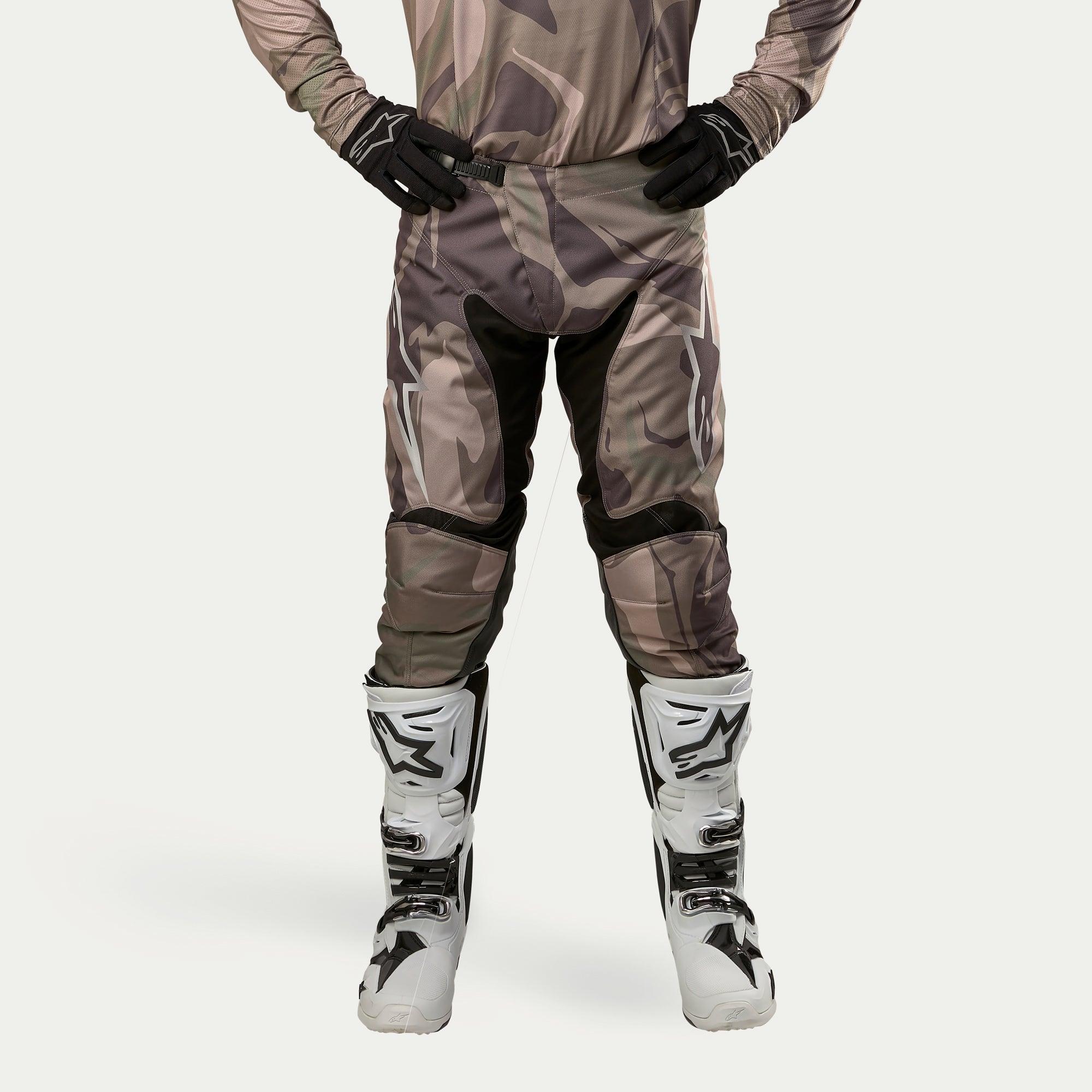 Alpinestars 2024 Racer Tactical Pants, calças de motocross, camuflado verde militar e castanho, com grandes logótipos Alpinestars brancos nas coxas e painéis pretos no interior das pernas, usadas com botas de motocross brancas e pretas, luvas pretas e camisola camuflada a condizer, vista frontal.